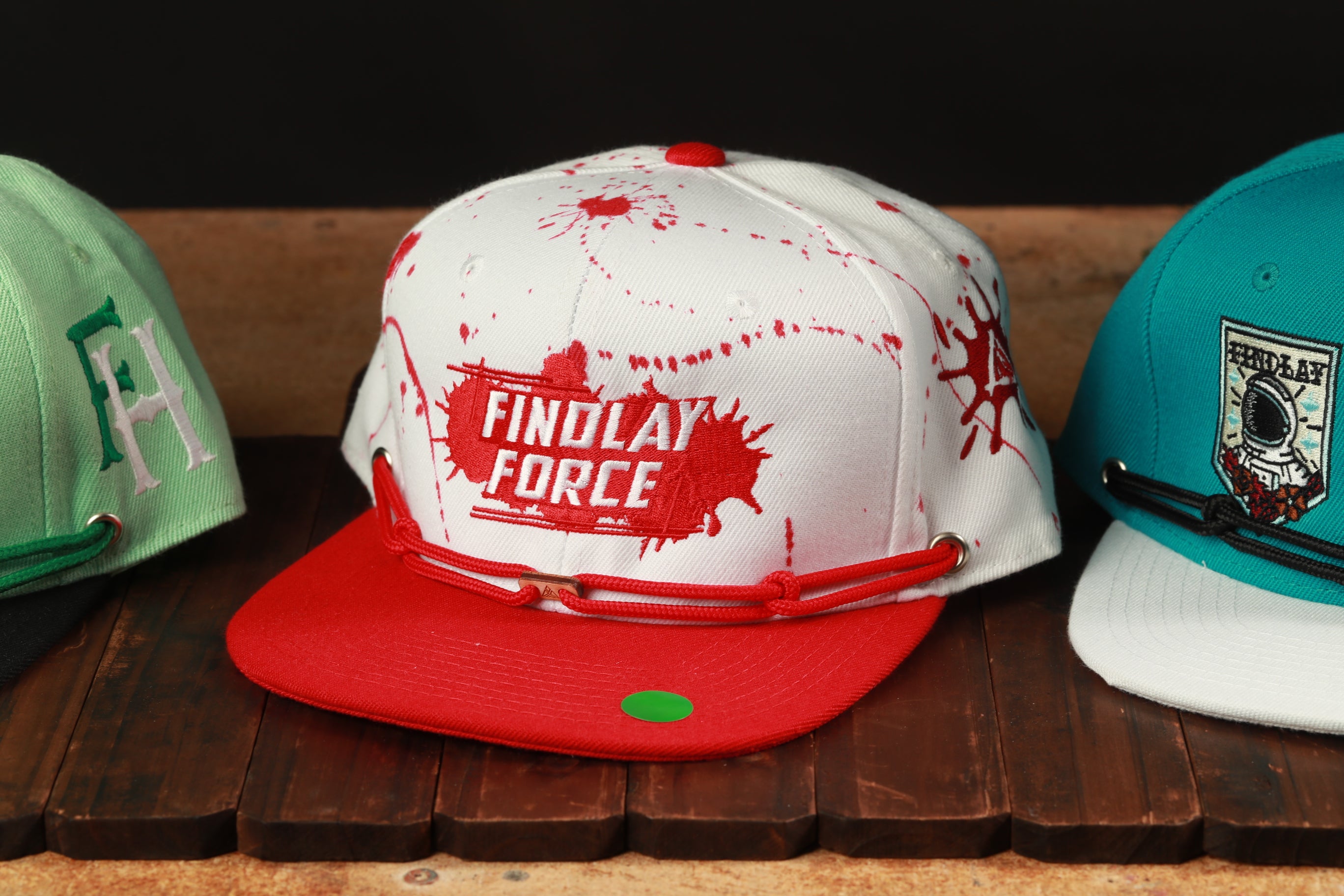 Three baseball caps are displayed on a wooden surface, with one prominently featuring the text "FINDOLAY FORCE" in red and white.