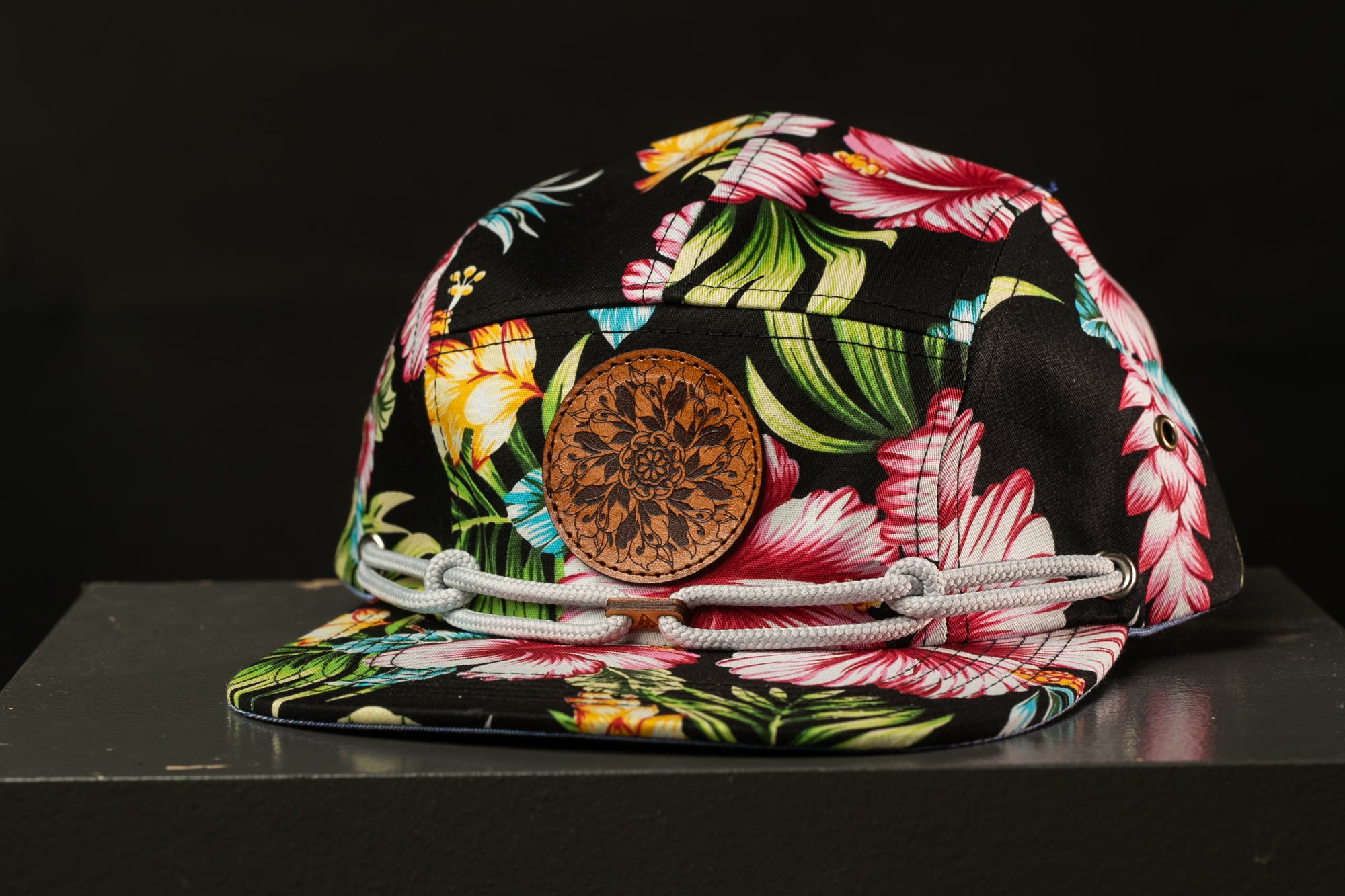 A colorful floral print cap with a leather patch and white straps, displayed on a black surface against a dark background.