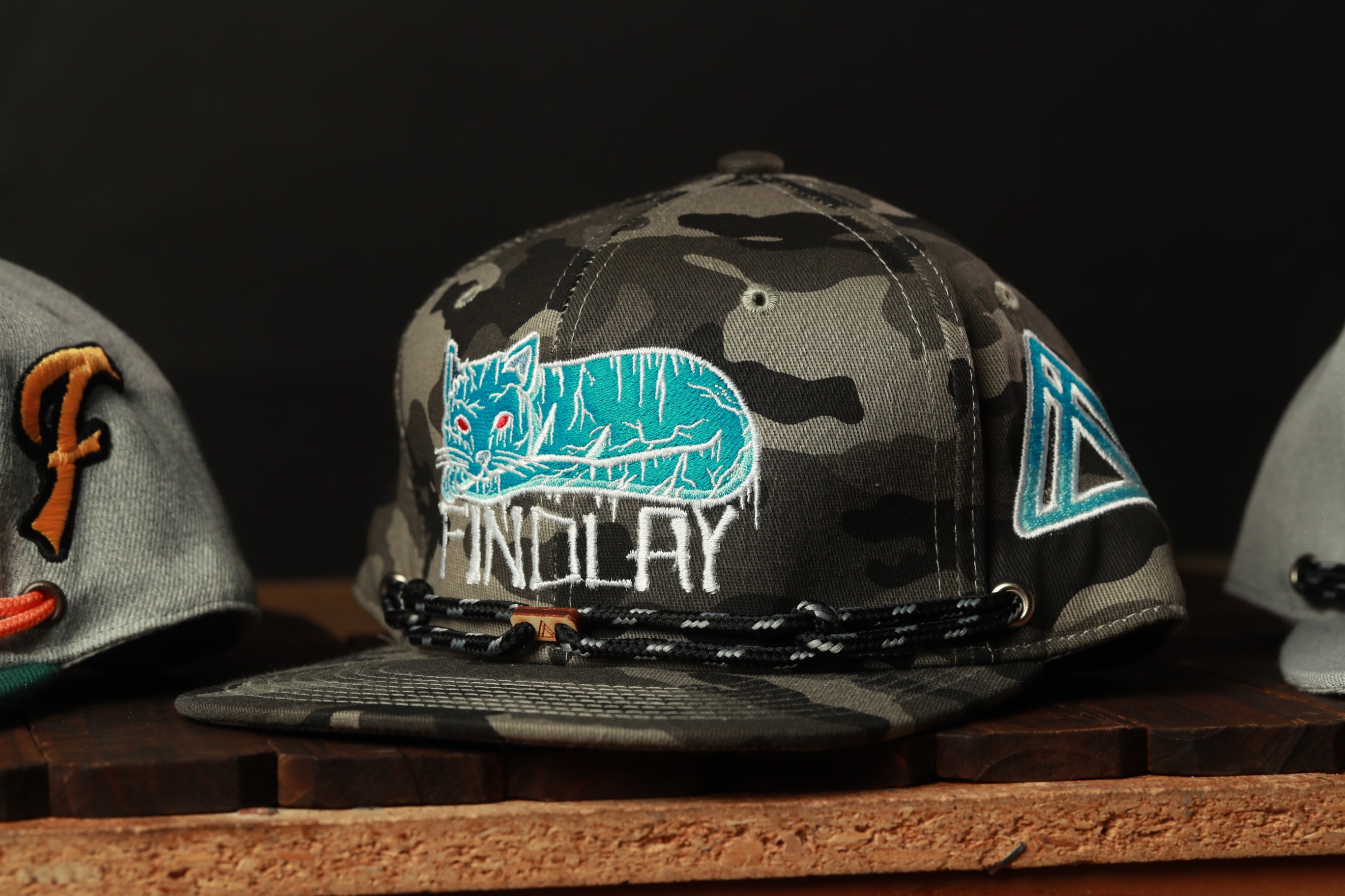 A camouflage baseball cap with a cat design and the word "FINDOLAY" printed on it, displayed on a wooden surface.