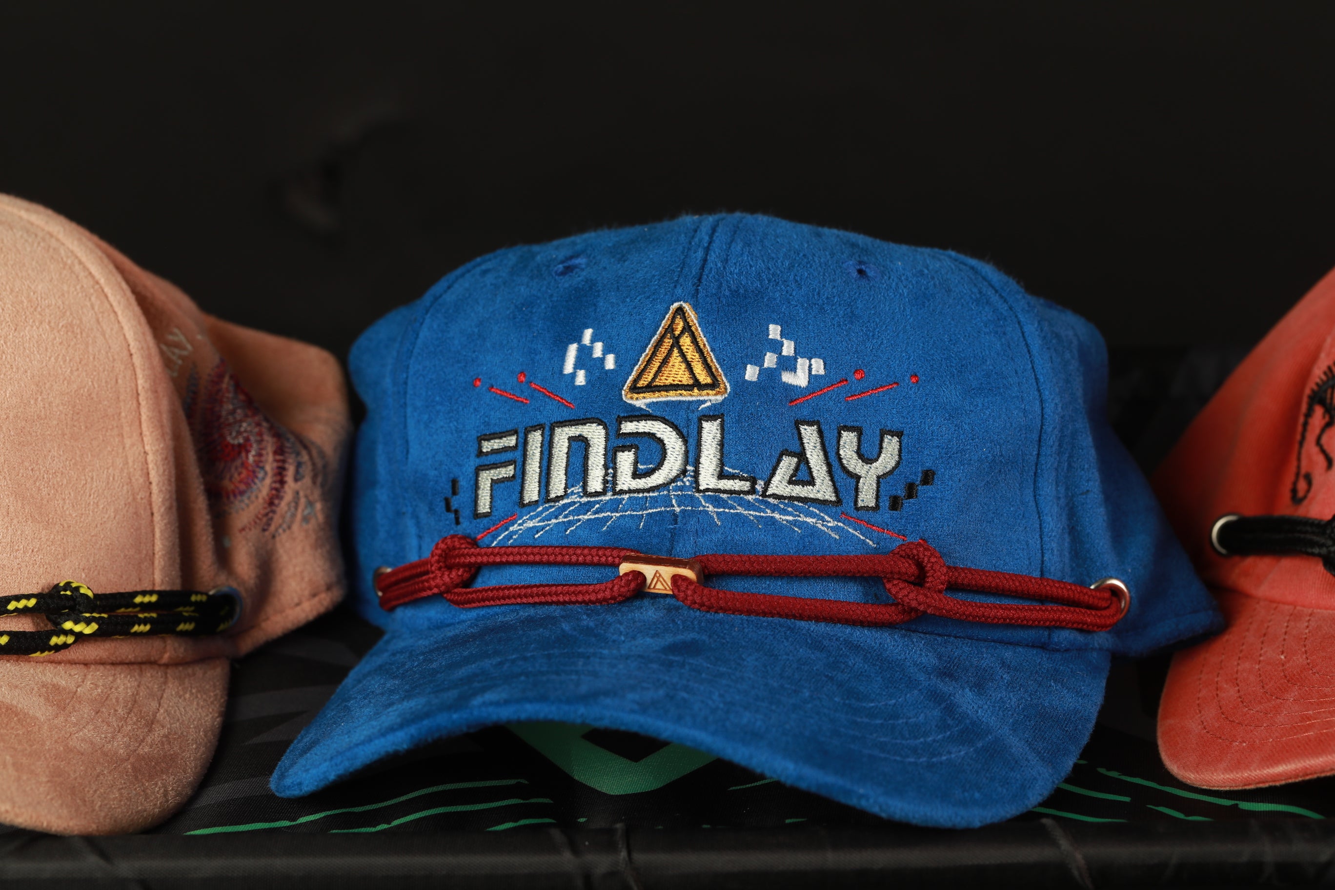 A blue baseball cap with the word "FINDlay" printed on it, along with a red string or strap, is displayed among other hats.