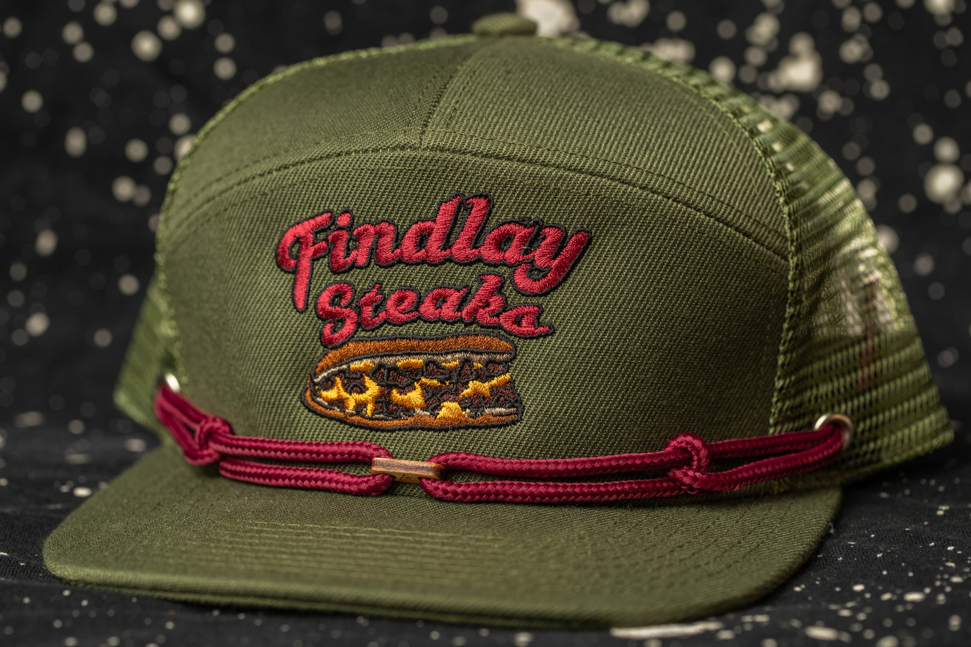 A green baseball cap with a red braided band and embroidered text on the front.