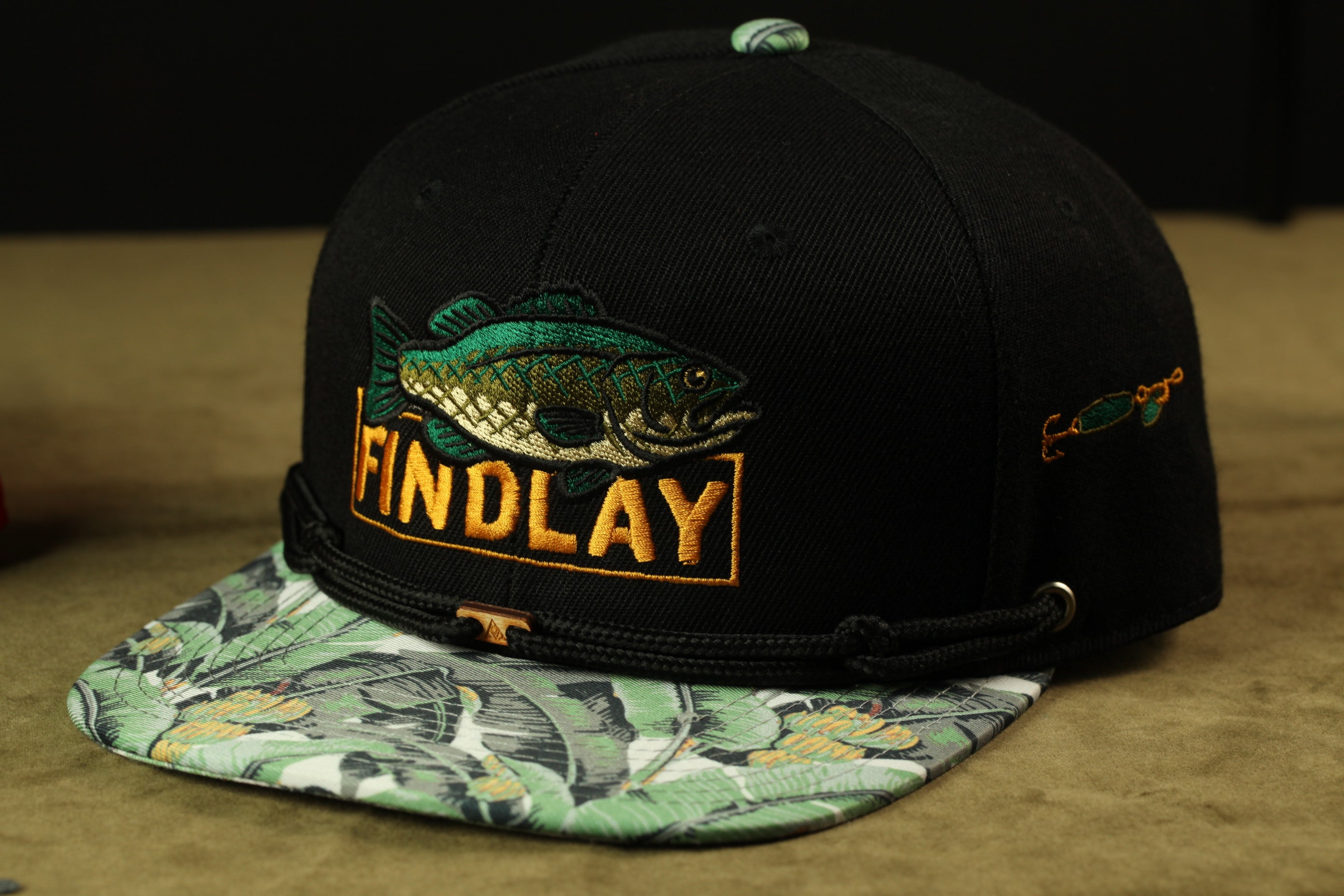 A black baseball cap with a fish embroidered on it, featuring the word "FINDLAY" in orange letters.