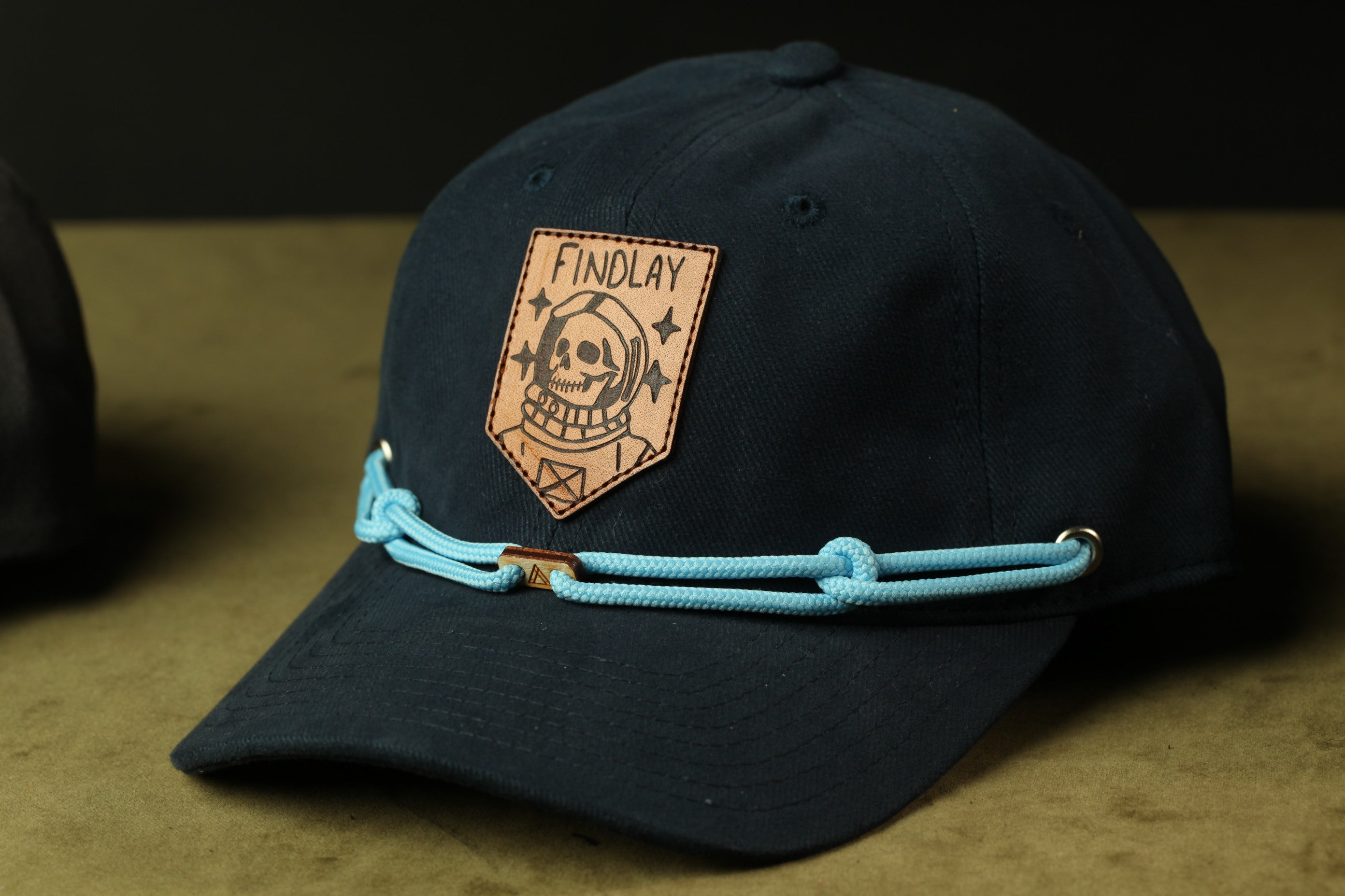 A black baseball cap with a leather patch on the front, featuring a skull and crossbones design, and a blue string or strap.