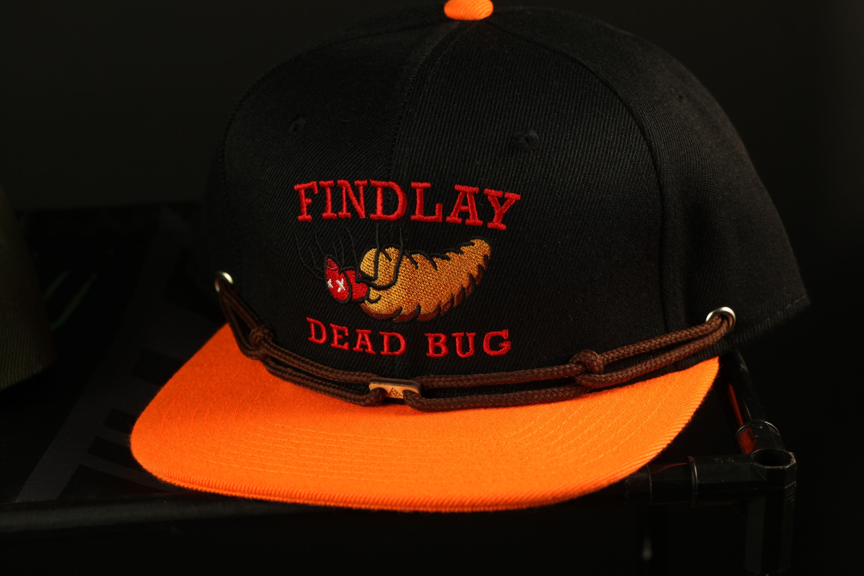 A black baseball cap with an orange brim, featuring a red and yellow design and the text "FIND LAY DEAD BUG" printed on it.