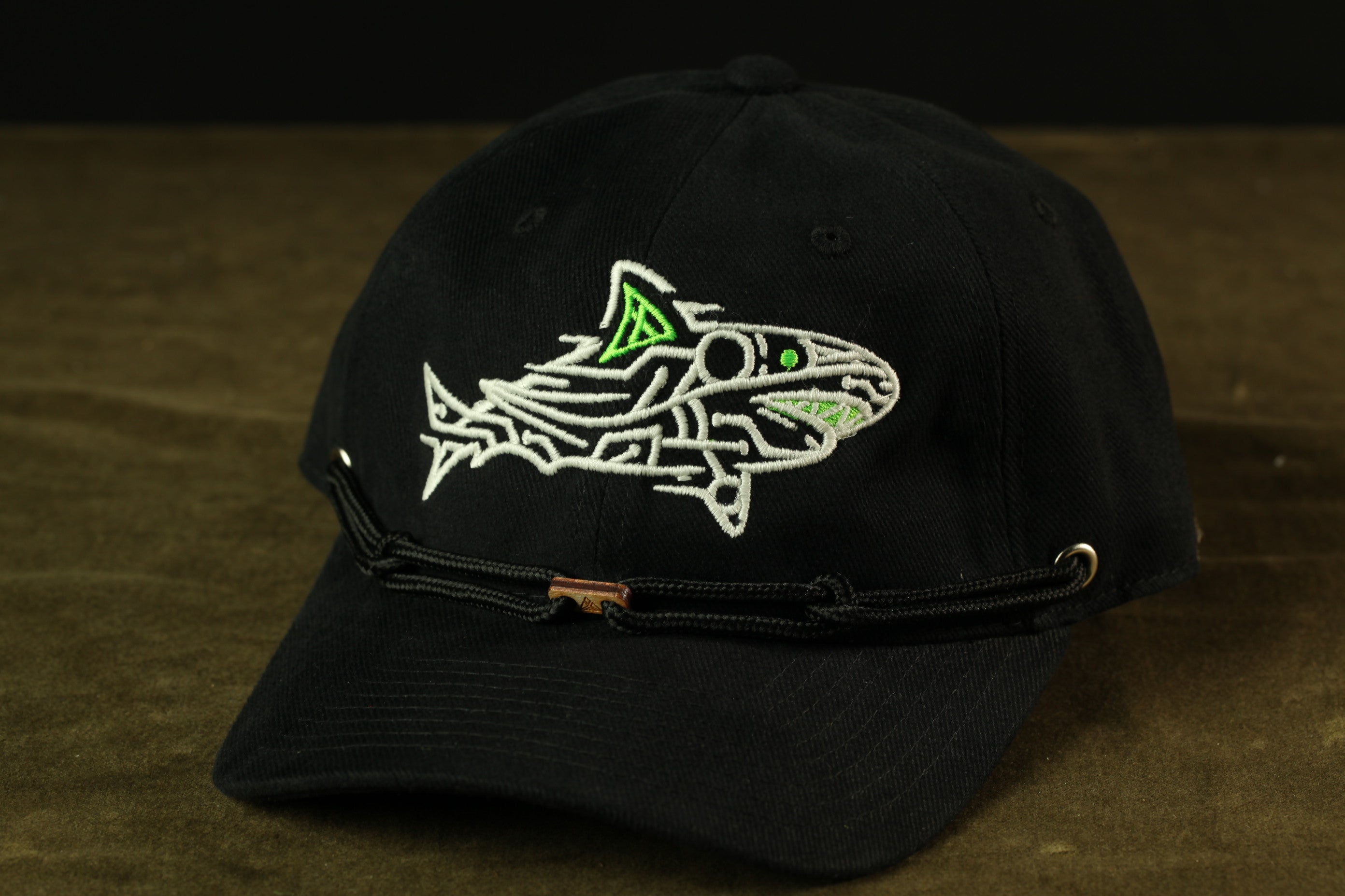A black baseball cap with a white fish embroidered on it, resting on a brown surface.