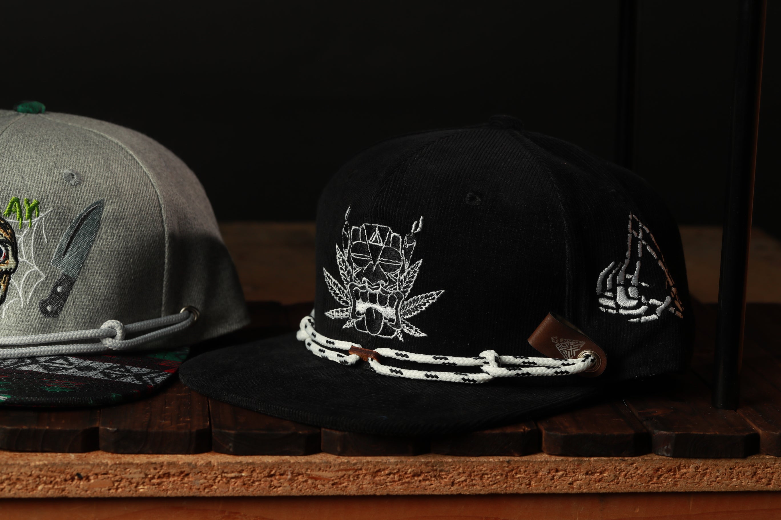 Two baseball caps are displayed on a wooden surface, one black and one gray, with various designs and patterns.