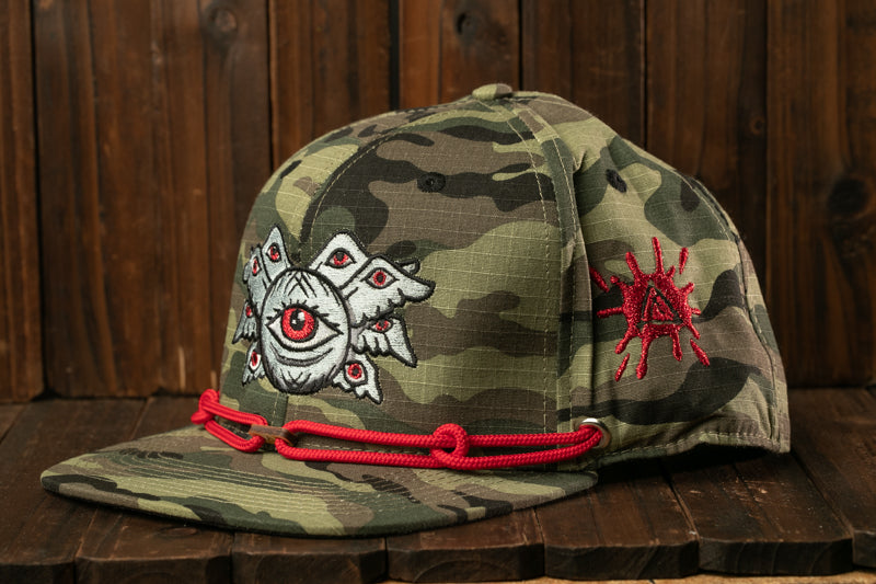 A camouflage baseball cap with a red string and an eye design on the front.