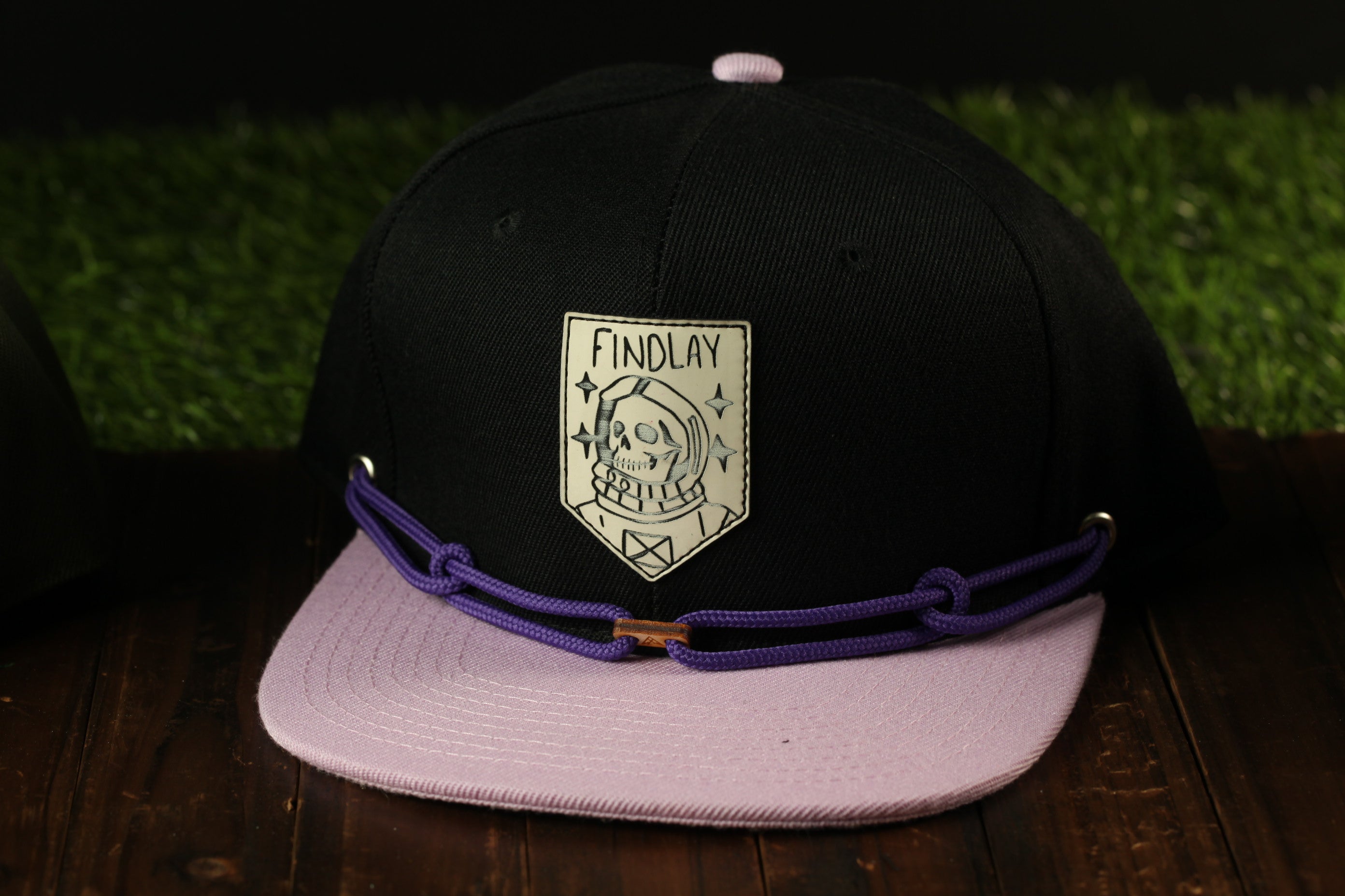 A black baseball cap with a purple band and a white patch on the front, featuring a graphic design.