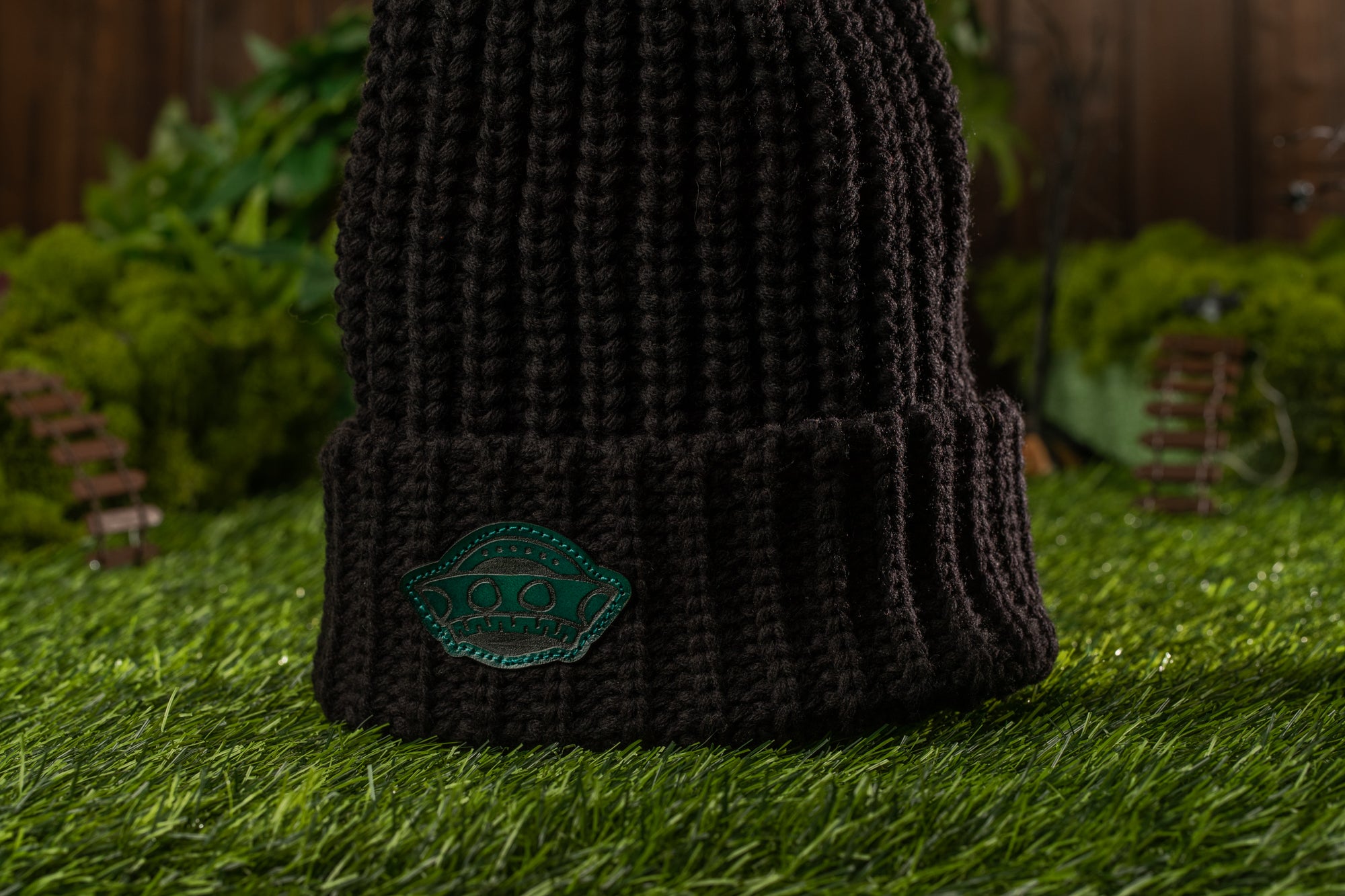 A dark knit beanie with a green patch on the front, placed on a grassy surface with a miniature garden in the background.