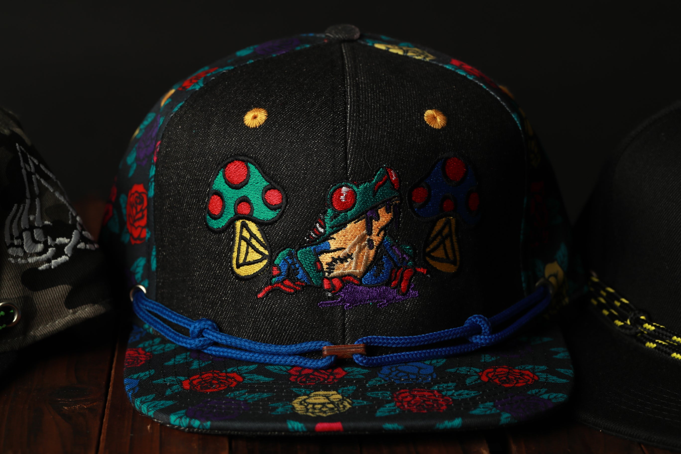 A colorful baseball cap with a frog design and mushrooms on it, sitting on a wooden surface.
