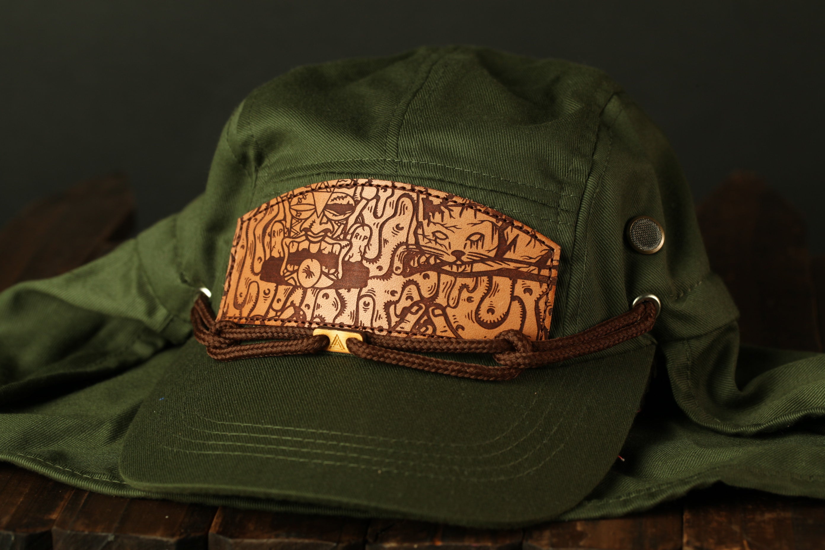 A green baseball cap with a leather patch featuring a cartoon-like design is displayed on a wooden surface.