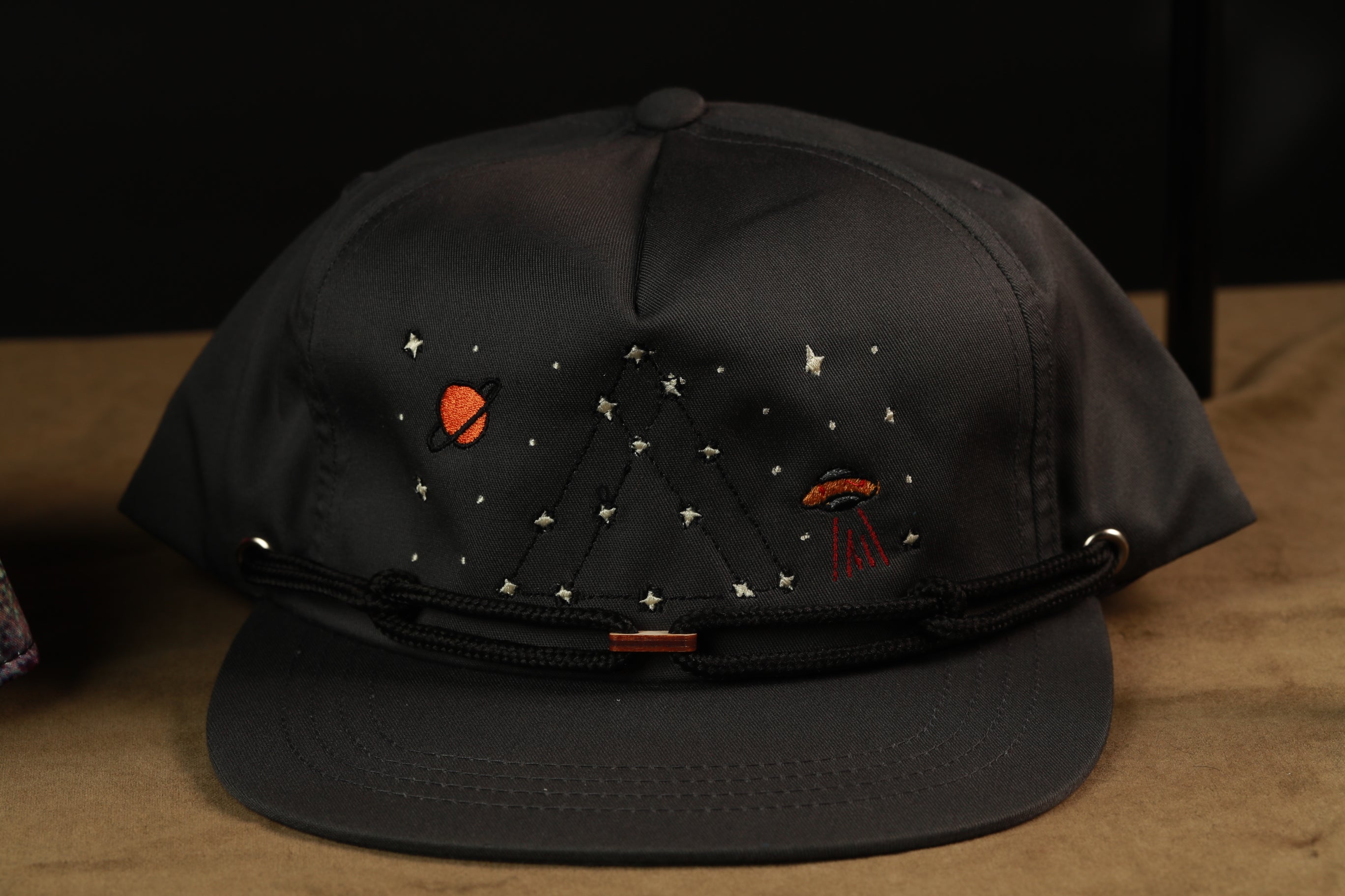 A black baseball cap with a space-themed design, including a planet, star, and alien figure, embroidered on the front.