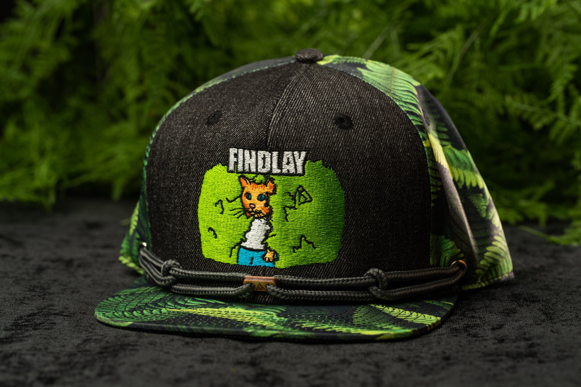 A black baseball cap with a green and black camouflage pattern, featuring a cartoon character on the front.
