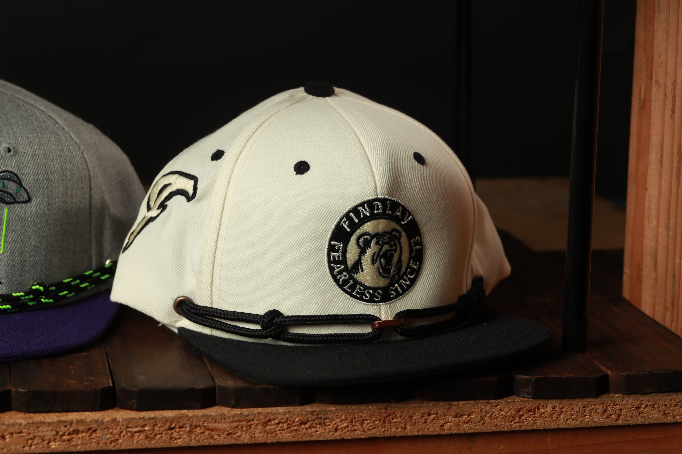 A white baseball cap with a black brim and a circular logo on the front, displayed on a wooden shelf.