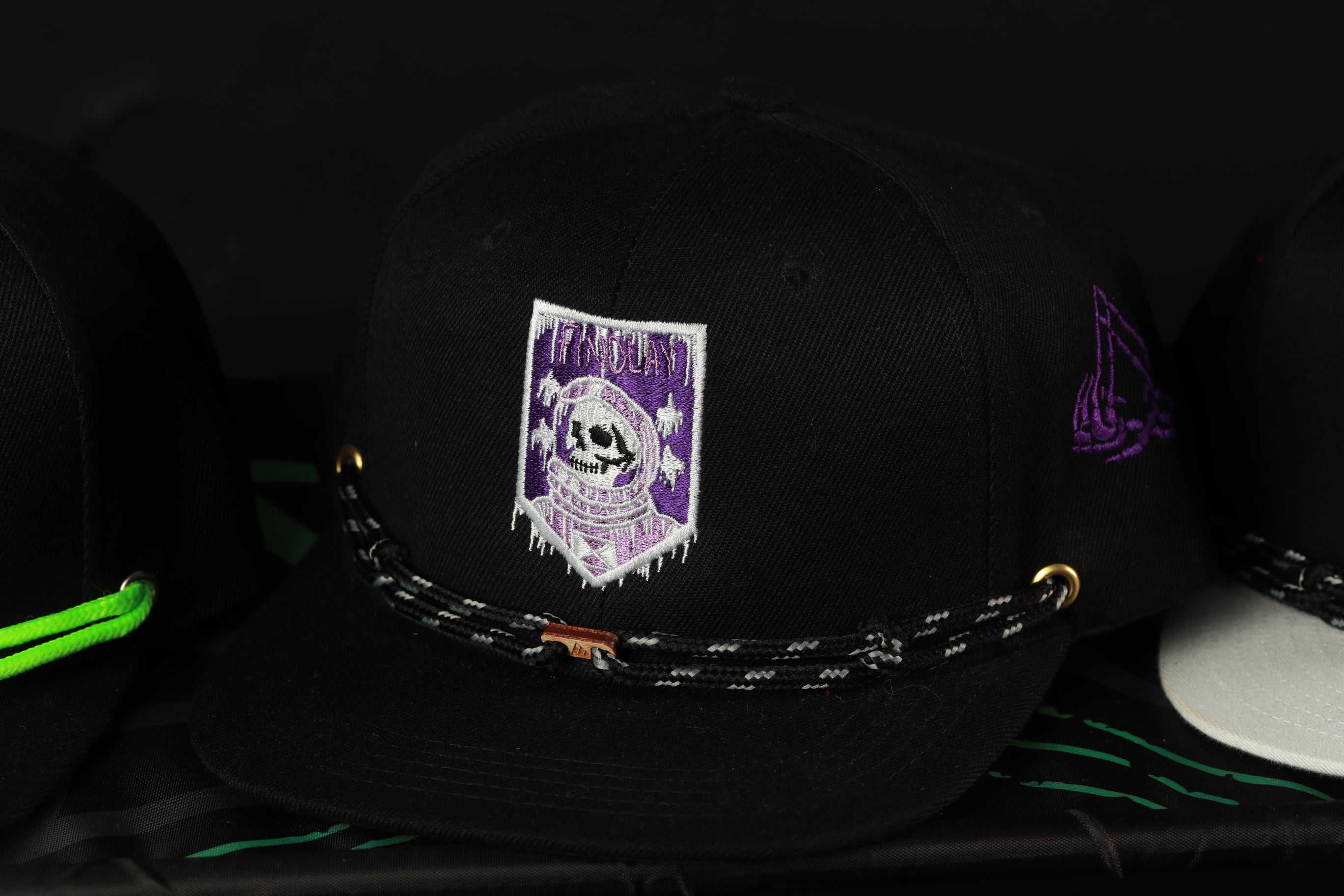 A black baseball cap with a purple and white skull embroidered on it, along with a black and white braided chain strap.