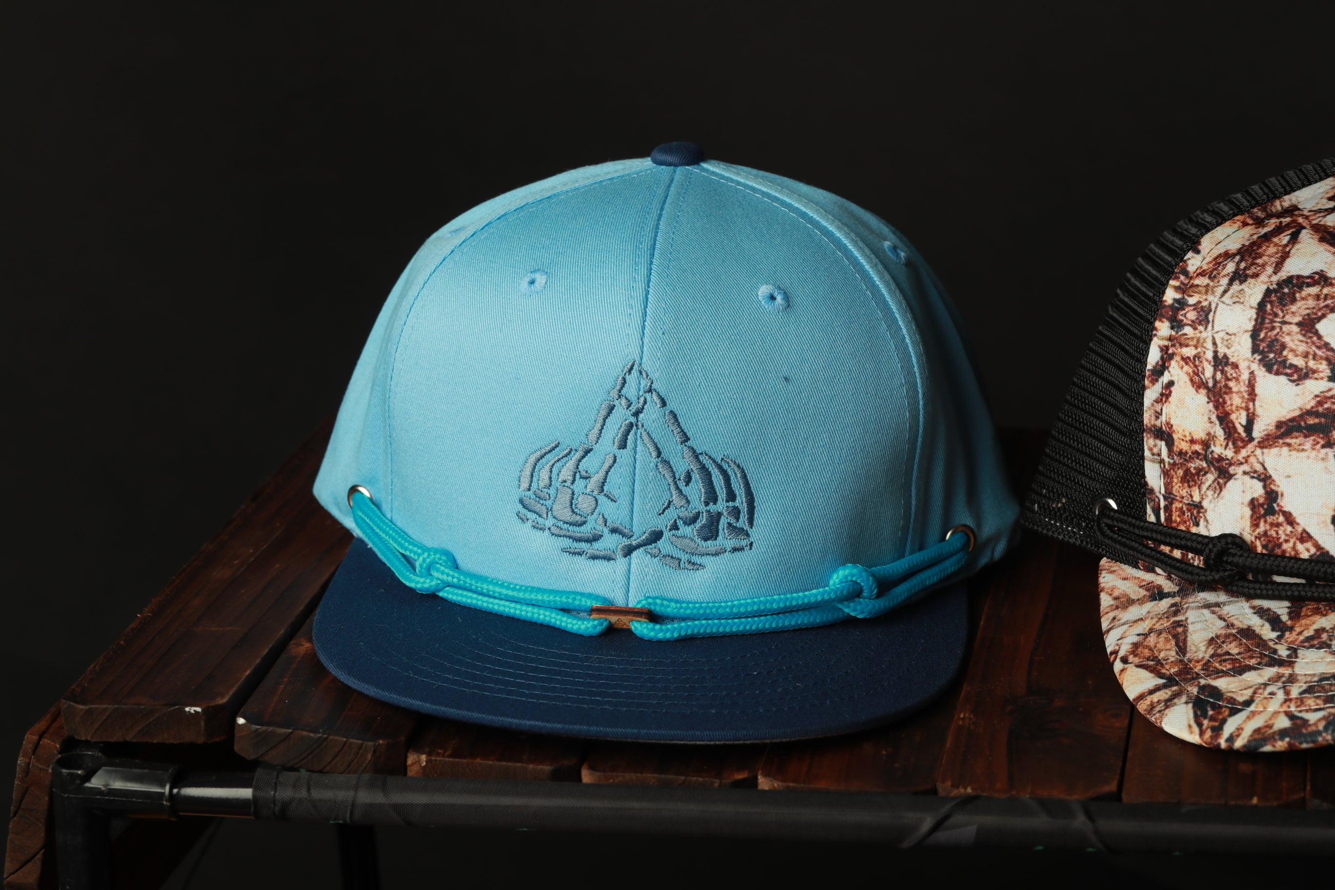 A blue baseball cap with a unique design on the front, placed on a wooden surface next to another cap.