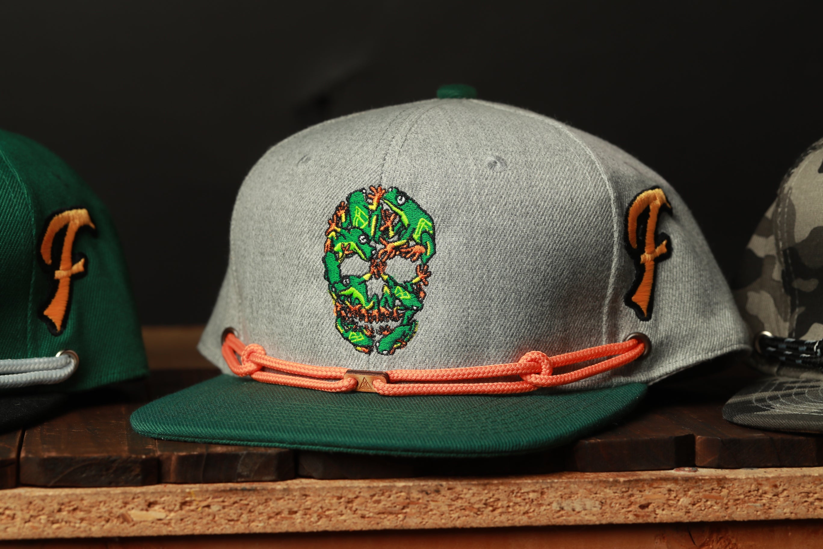 A gray baseball cap with a skull design and orange straps is displayed on a wooden surface.