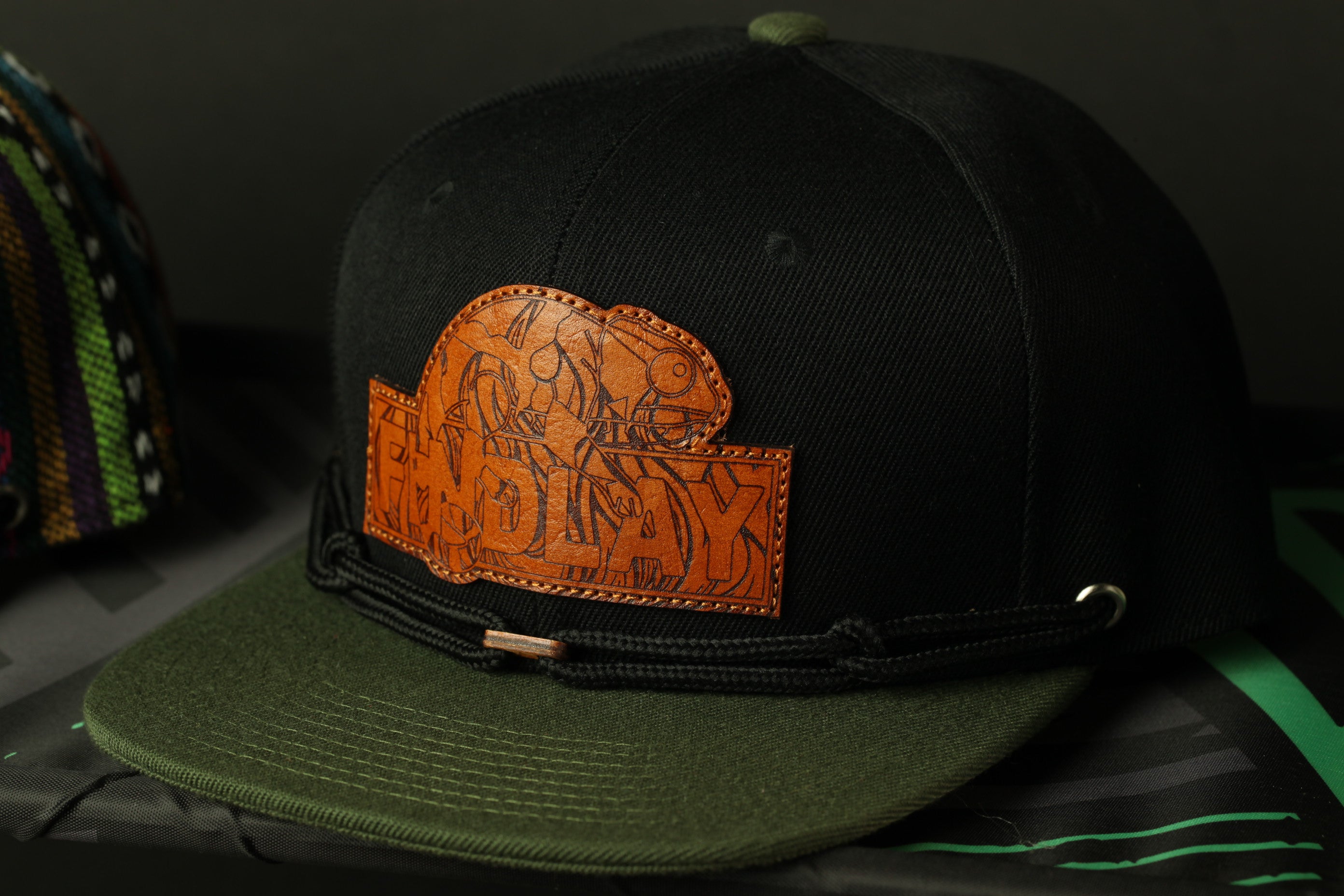 A black baseball cap with a leather patch on the front, featuring an orange design.