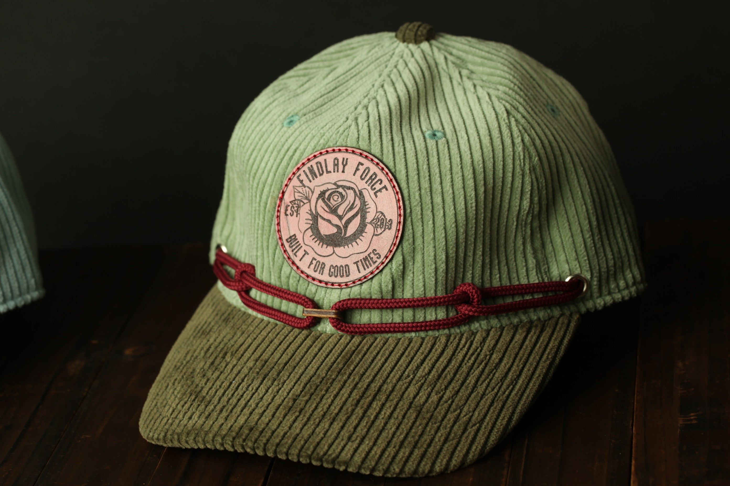 A green baseball cap with a red ribbon and a circular patch on the front, sitting on a wooden surface.