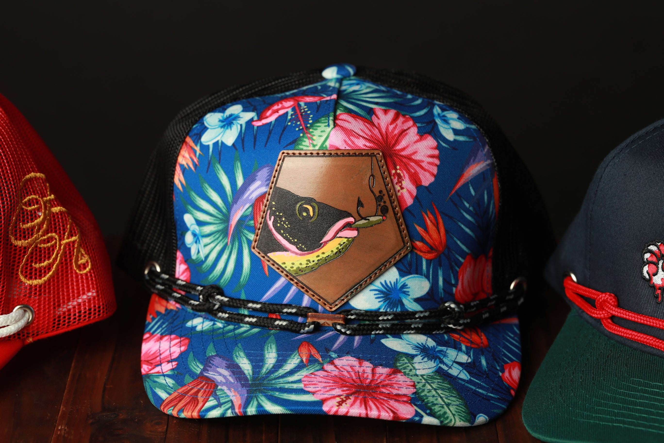 A colorful floral-patterned baseball cap with a brown leather patch featuring a fish design, displayed on a wooden surface.