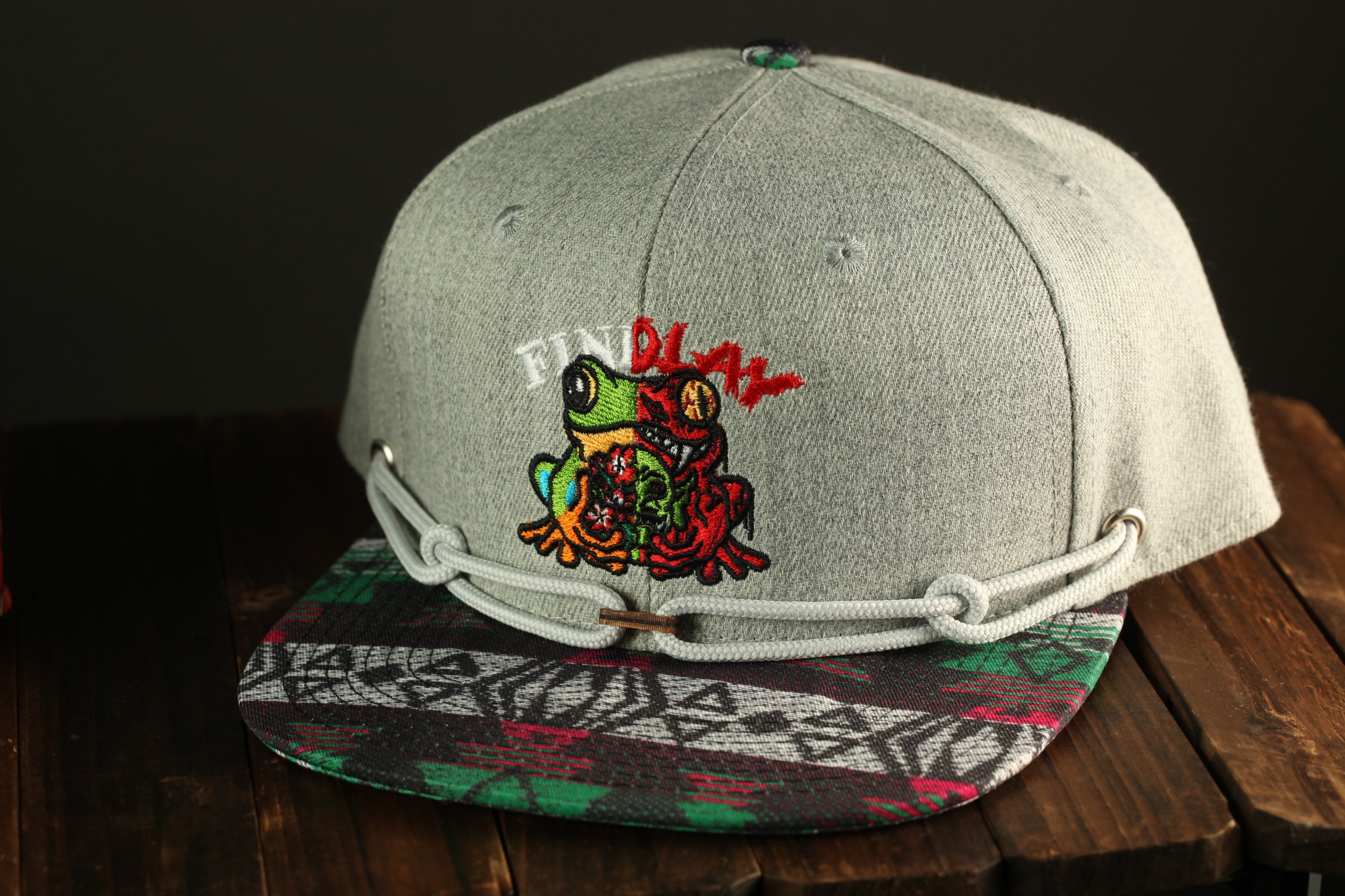 A gray baseball cap with a colorful frog embroidered on it, sitting on a wooden surface.