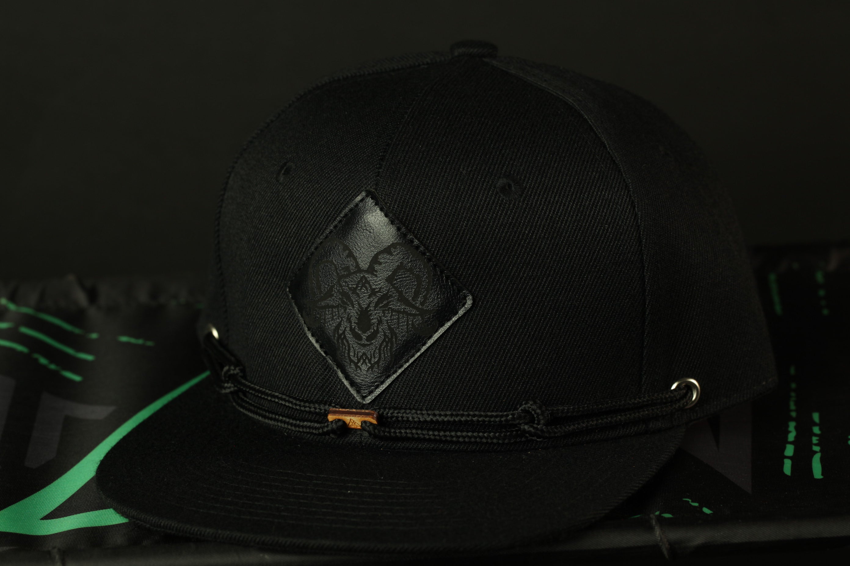A black baseball cap with a distinctive logo on the front, resting on a surface with a green and black patterned background.