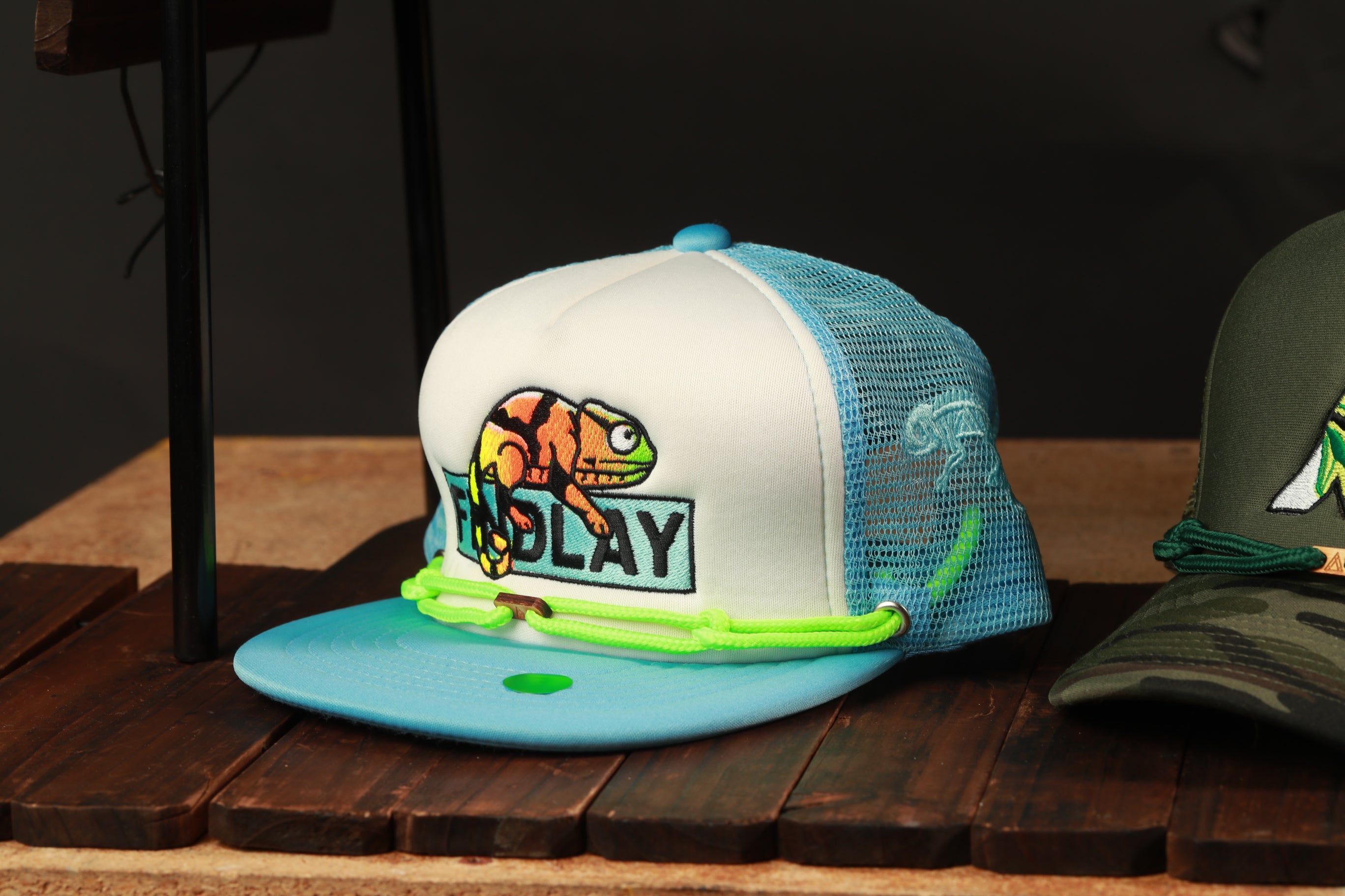 A white baseball cap with a colorful frog design and the word "PLAY" printed on it, sitting on a wooden surface.