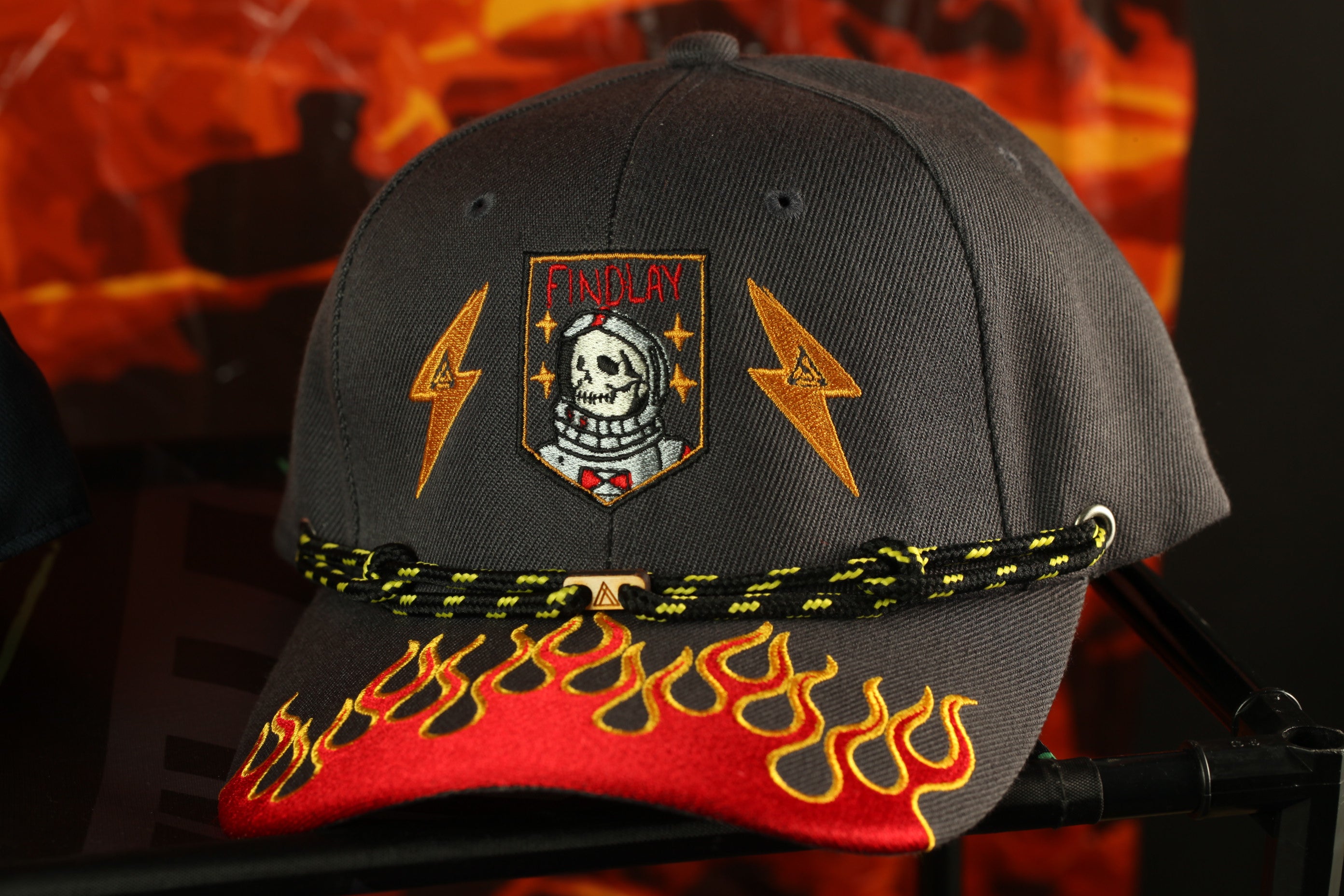 A black baseball cap with a flame-designed brim and a skull-emblazoned patch on the front, set against a fiery background.