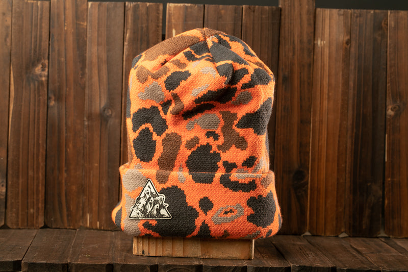 An orange and black camouflage beanie with a triangular logo on the front, displayed on a wooden surface against a wooden background.