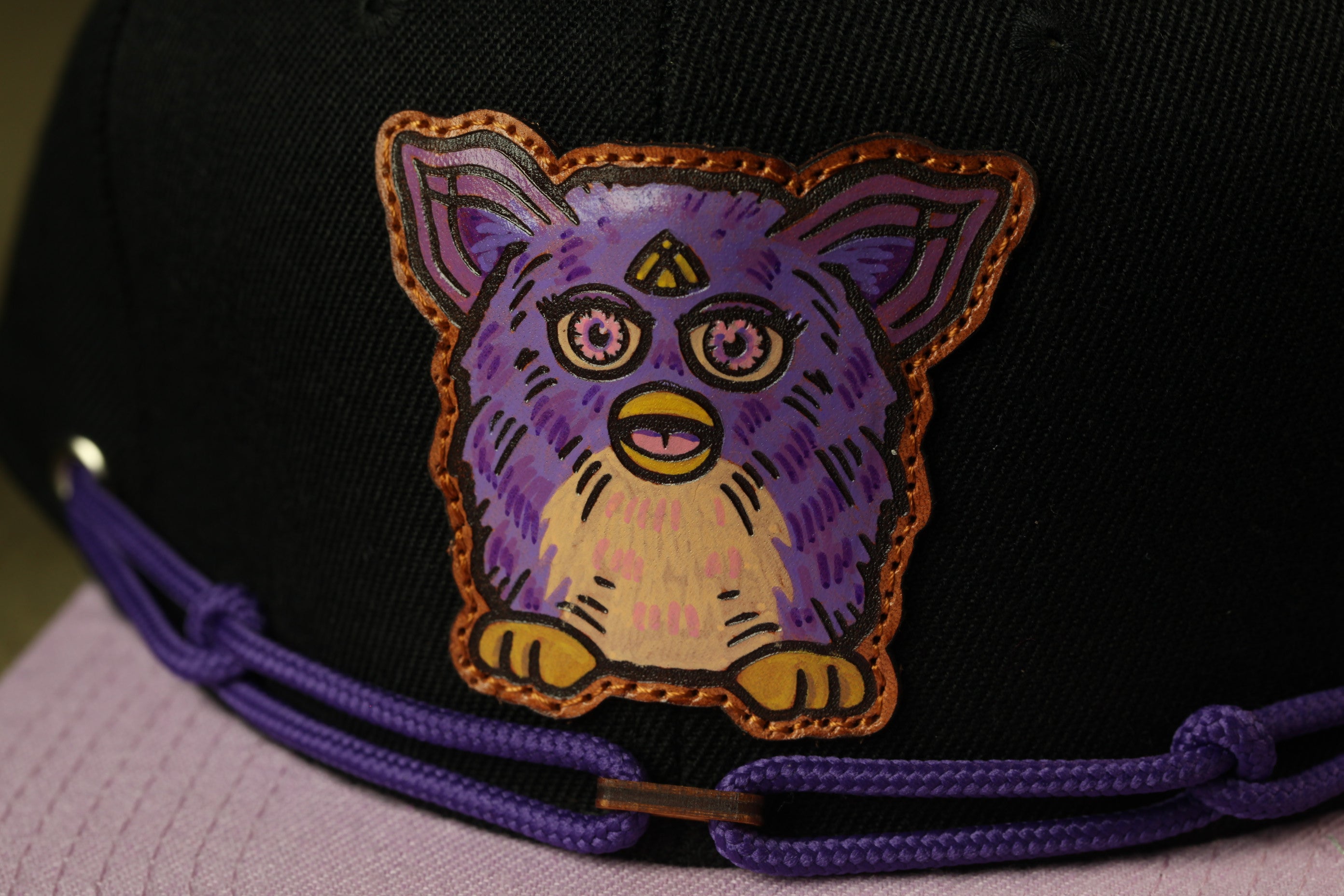 A close-up of a black baseball cap with a purple and yellow cartoon character design on the front.