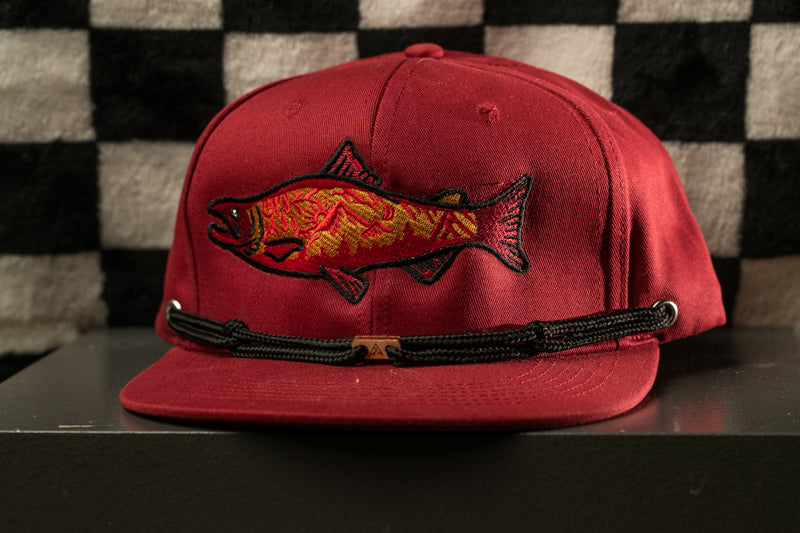 A red baseball cap with a fish embroidered on the front, displayed on a black surface against a checkered background.
