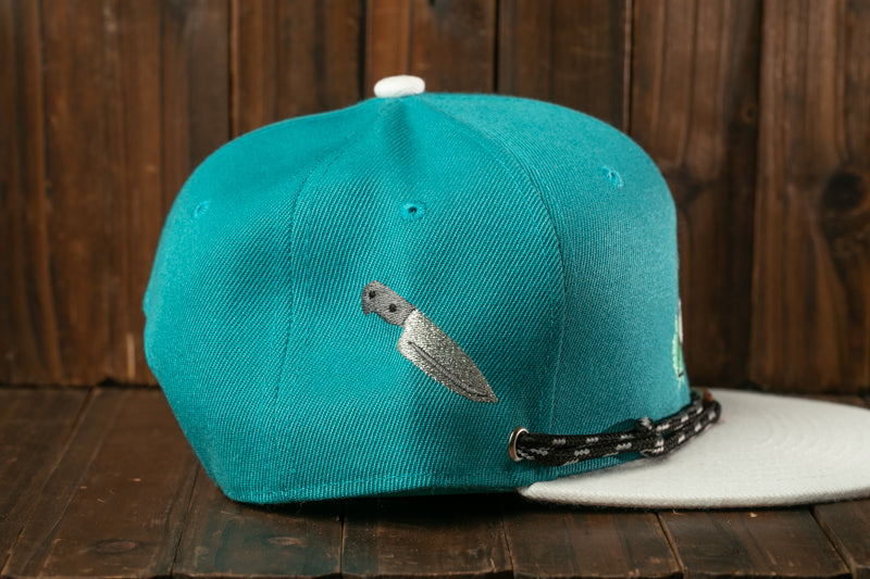 A turquoise baseball cap with a knife embroidered on it, placed on a wooden surface.