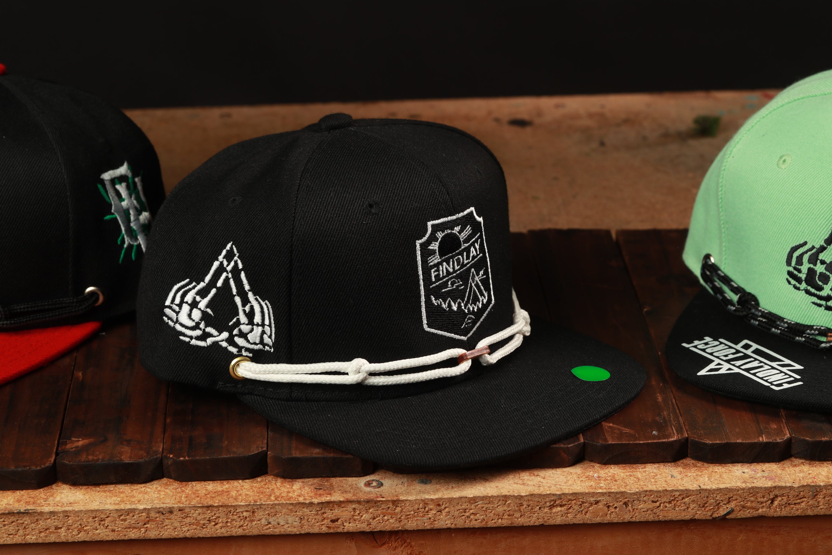 A black baseball cap with white text and graphics is displayed on a wooden surface, along with another green baseball cap.