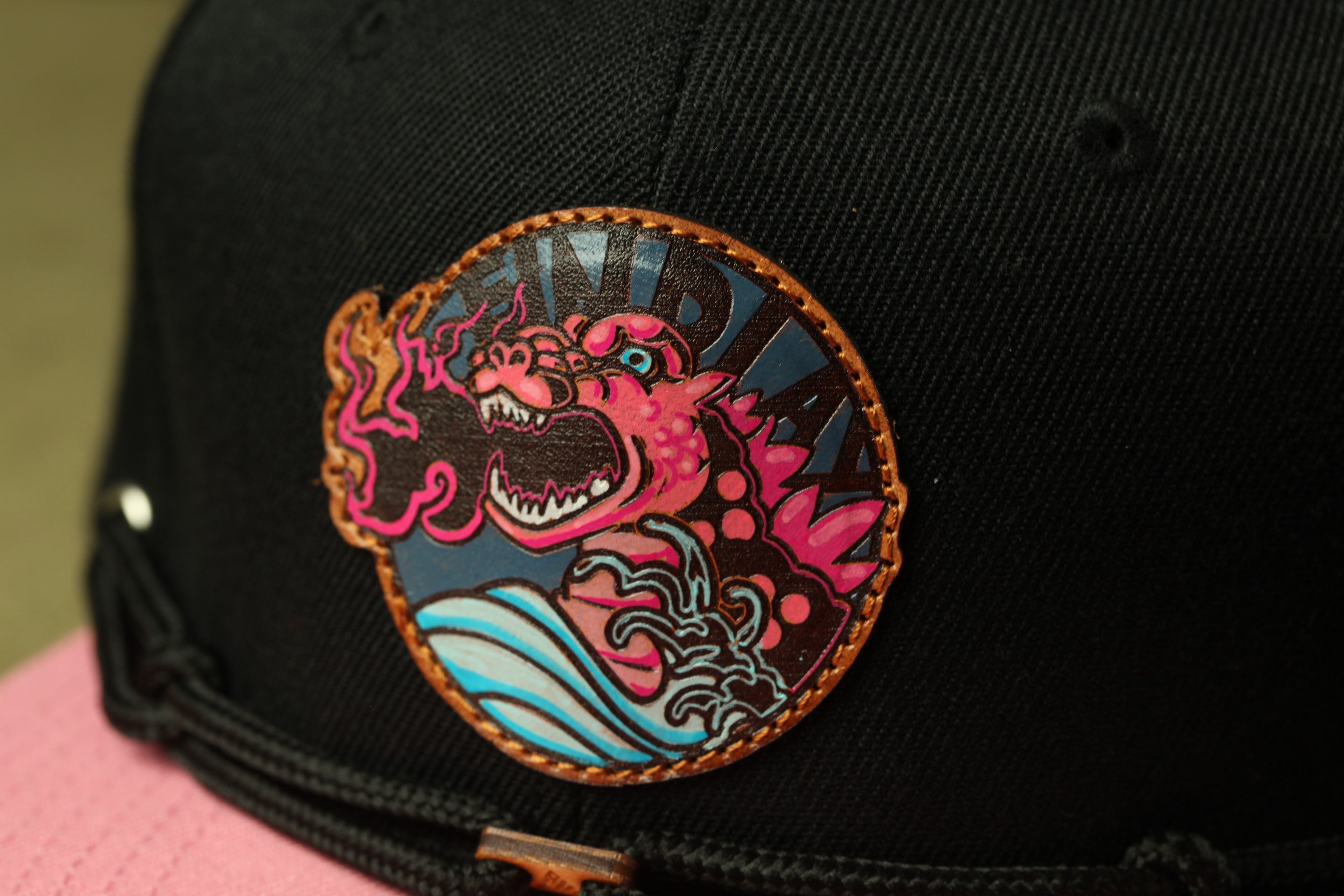 A close-up view of a black baseball cap with a colorful, cartoonish design featuring a large, fierce-looking creature with a pink and black color scheme.