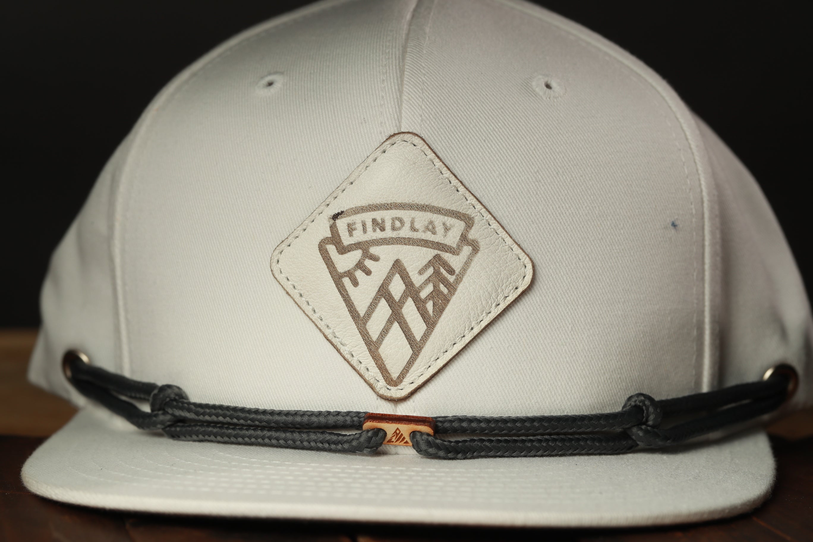 A close-up view of a white baseball cap with a leather patch on the front, featuring a logo and a mountain design.