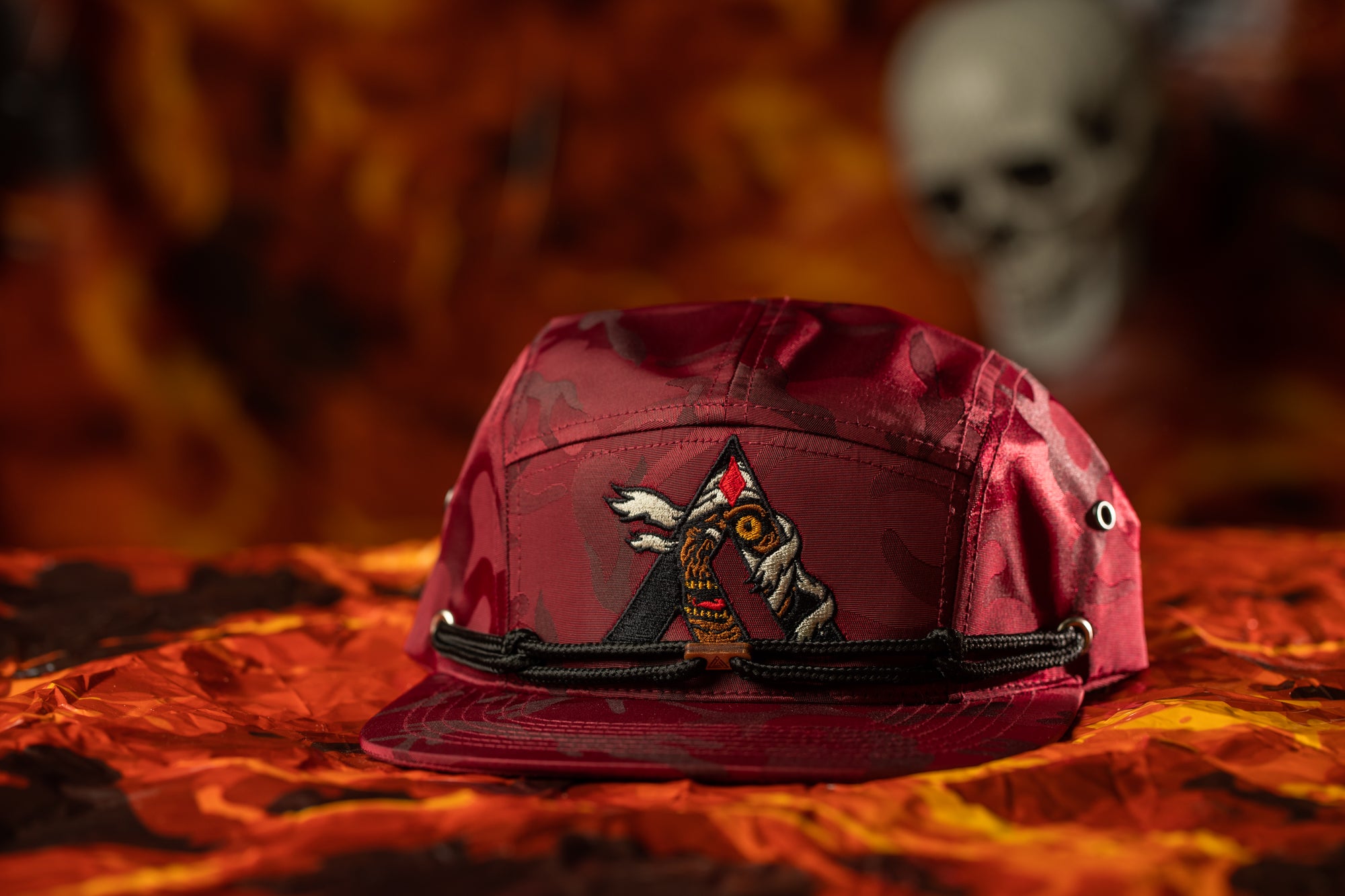 A maroon baseball cap with a skull design on the front, placed on a red and orange patterned surface.