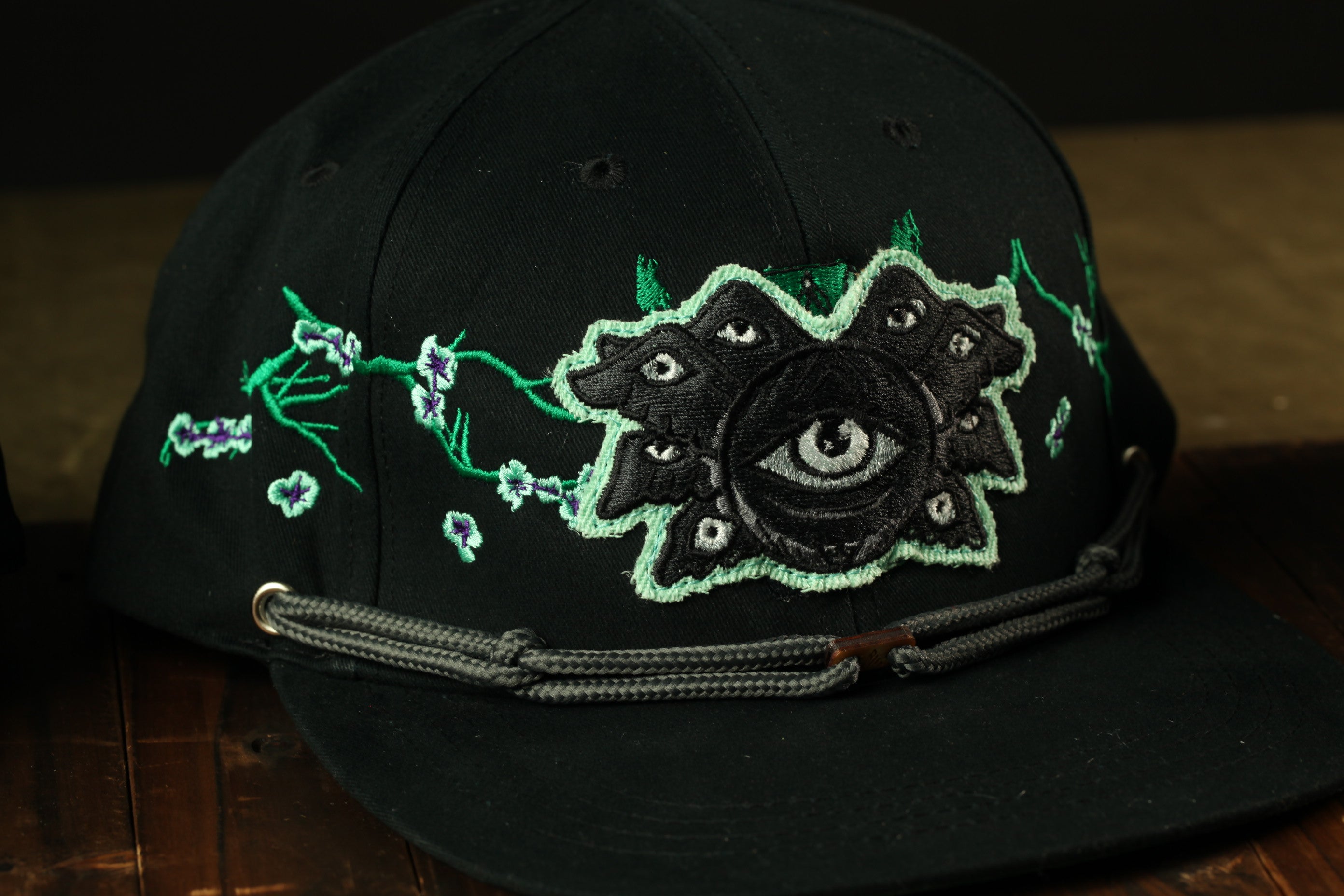 A black baseball cap with a unique embroidered design on the front, featuring a large eye surrounded by smaller eyes and floral elements.