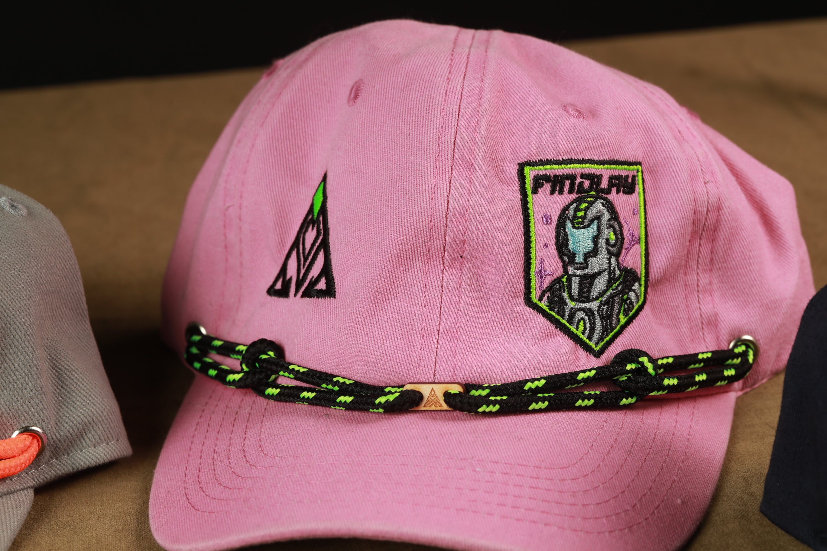 A pink baseball cap with a black and green braided strap and a patch featuring a robot or robot-like figure.