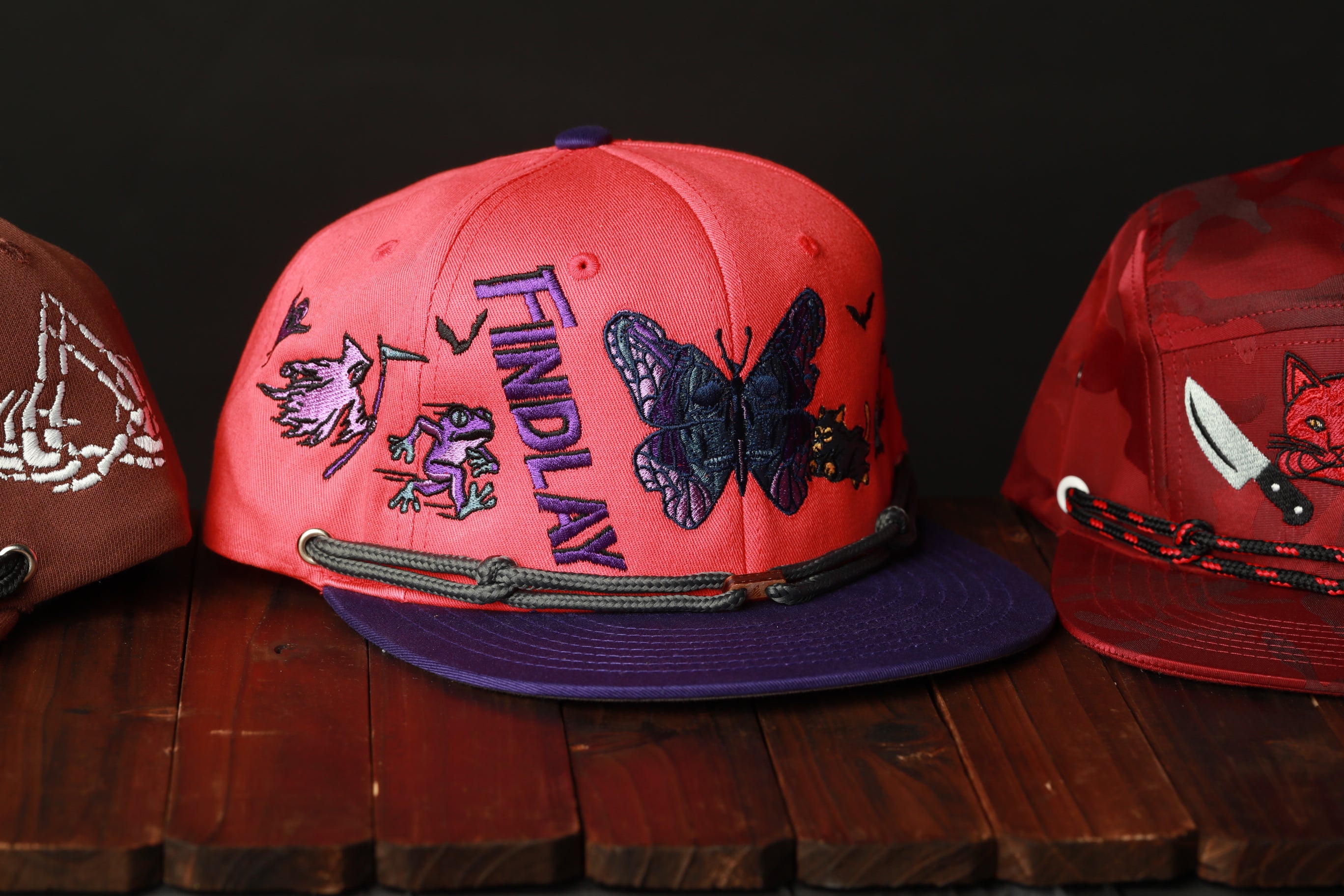 A red baseball cap with a purple brim and a butterfly design, along with other hats, are displayed on a wooden surface.