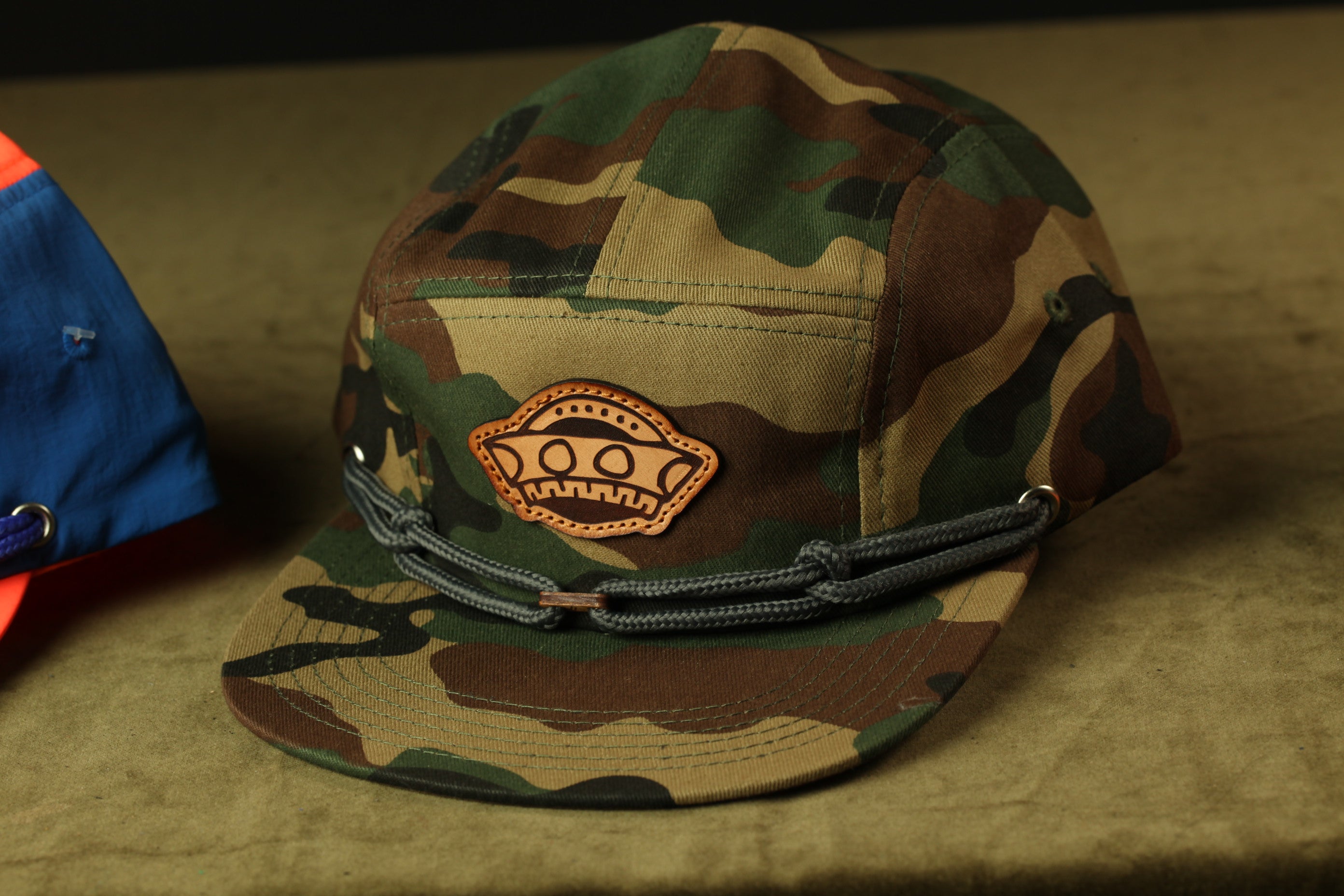 A camouflage baseball cap with a leather patch on the front, sitting on a brown surface.