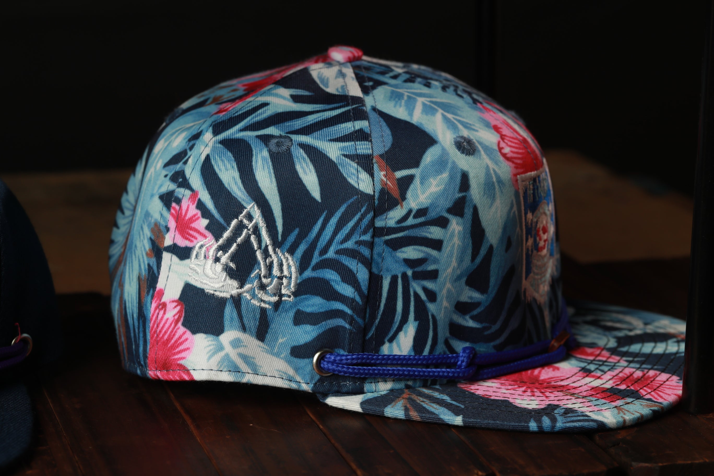 A colorful floral baseball cap with a blue brim and a blue strap, displayed on a wooden surface.