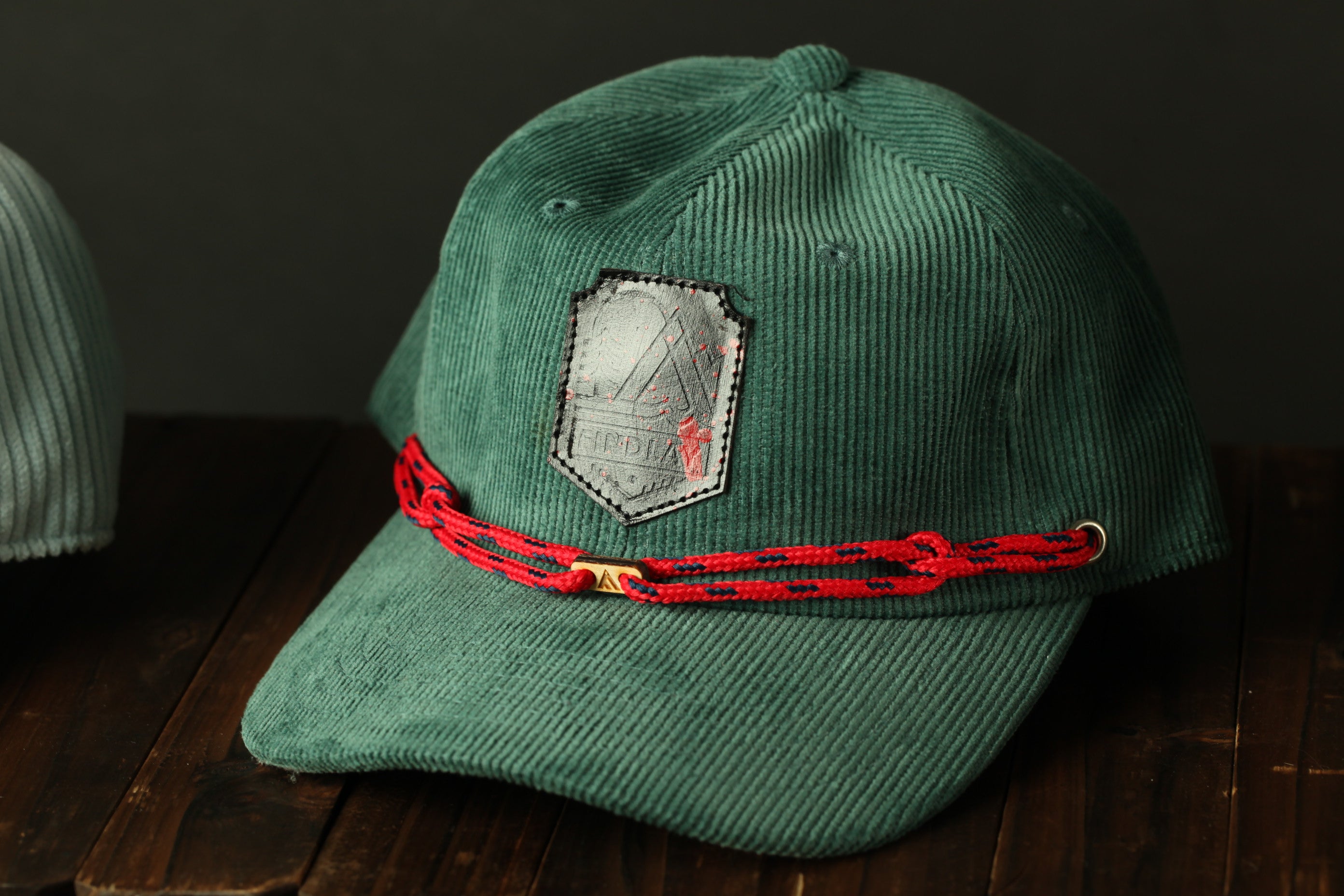 A green baseball cap with a red braided strap and a patch on the front, placed on a wooden surface.