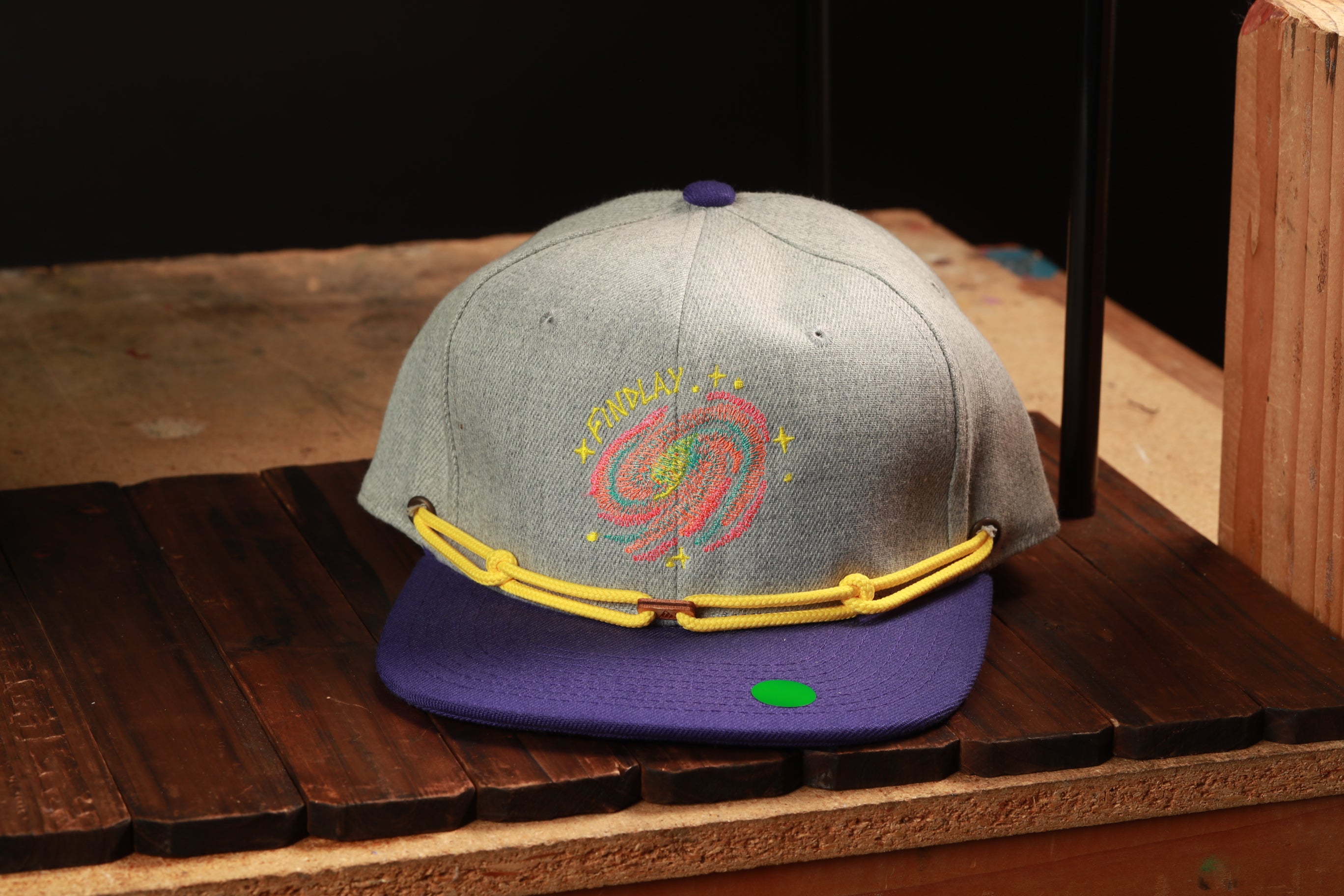 A gray baseball cap with a purple brim and yellow string, featuring a colorful spiral design on the front, is displayed on a wooden surface.