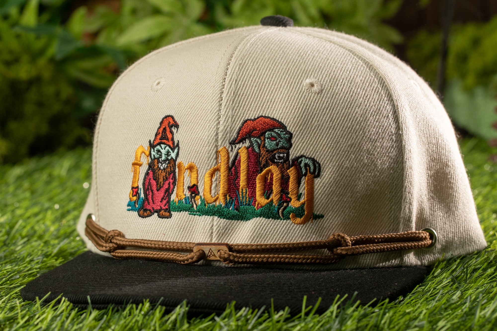 A baseball cap with a cartoonish design on it, featuring a gnome and a dragon-like creature, is placed on a grassy surface.