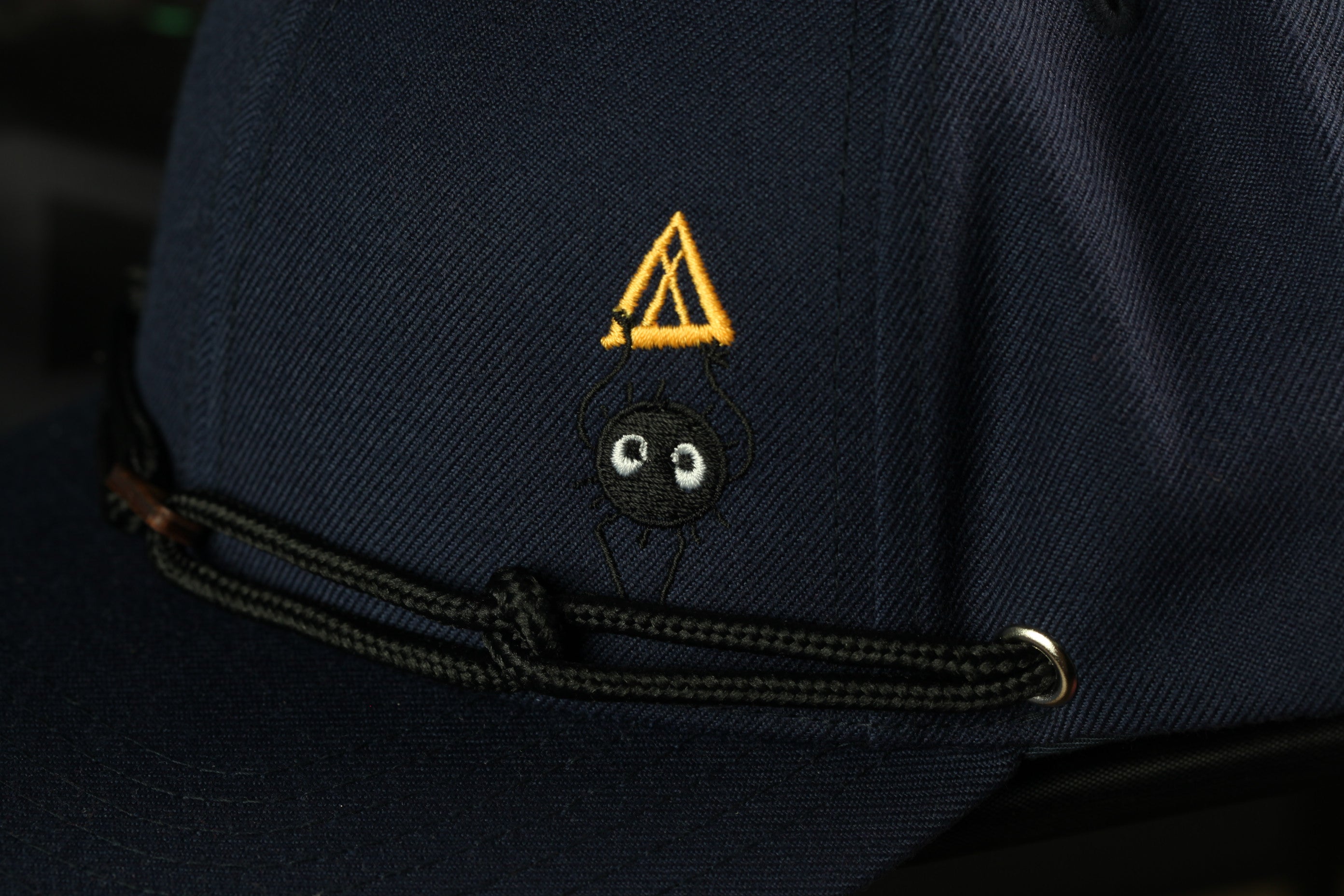 A close-up view of a navy blue baseball cap with a small black spider embroidered on the front.