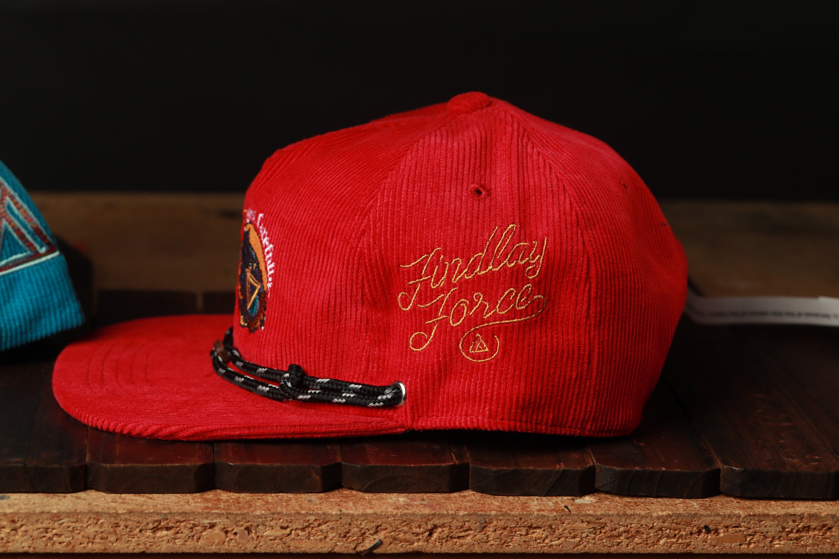 A red baseball cap with a black braided strap and embroidered text on the front, sitting on a wooden surface.