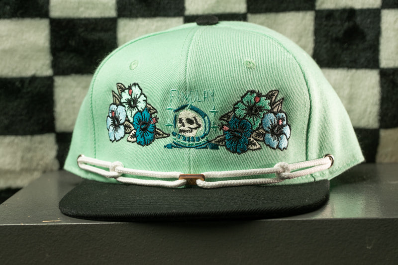 A mint green baseball cap with floral embroidery and a skull design, displayed on a black surface against a checkered background.