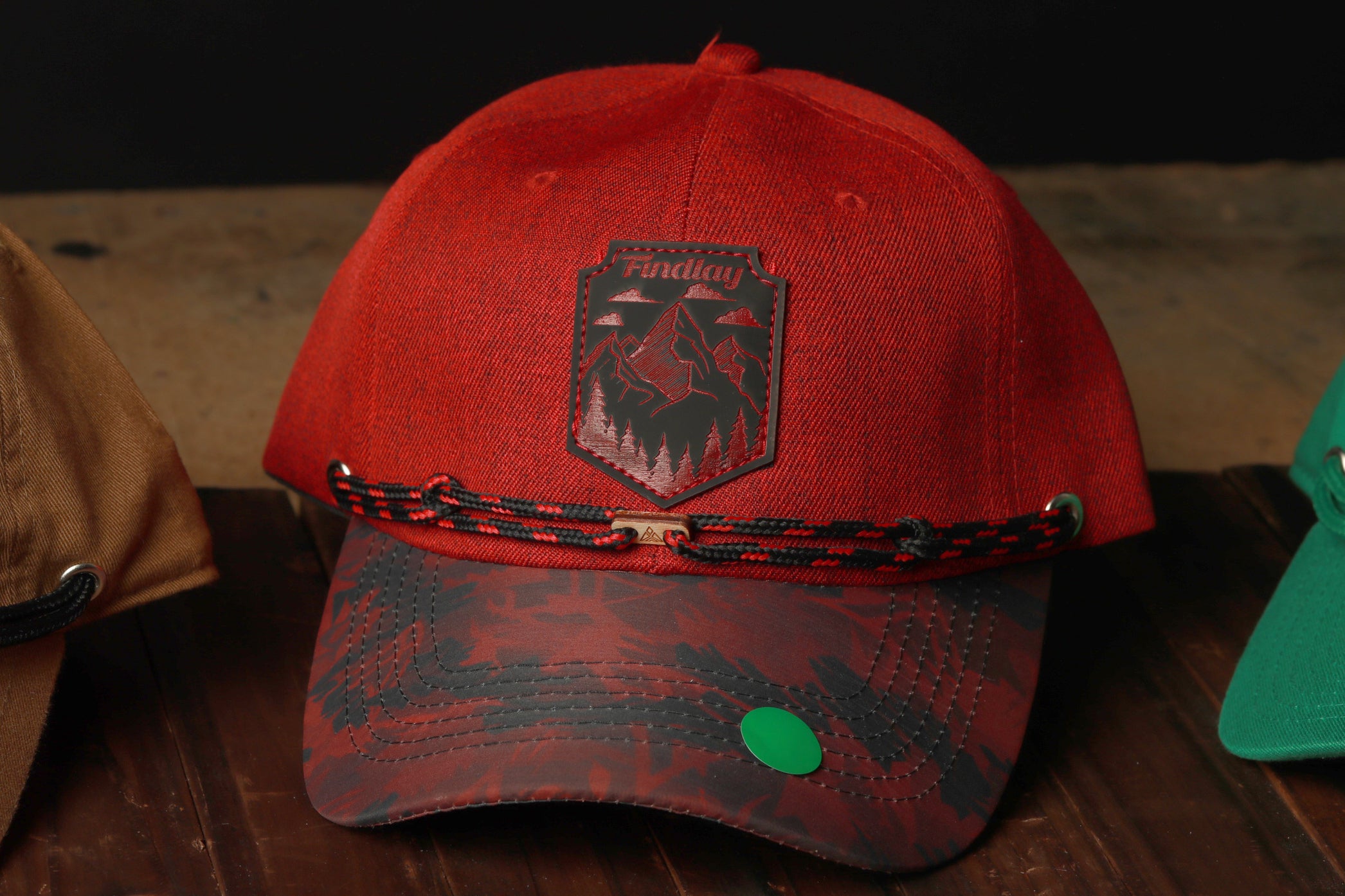 A red baseball cap with a mountain-themed patch and a black and red braided strap is displayed on a wooden surface.