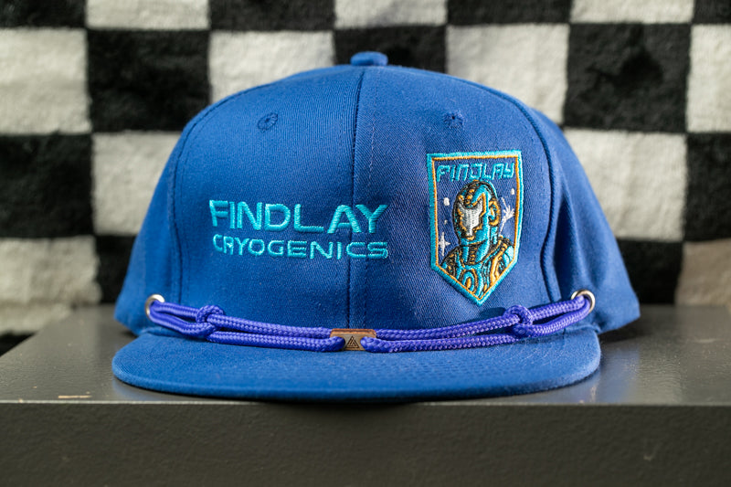 A blue baseball cap with the text "FINDLAY CRYOGENICS" and a small emblem on the front, placed on a black surface against a checkered background.