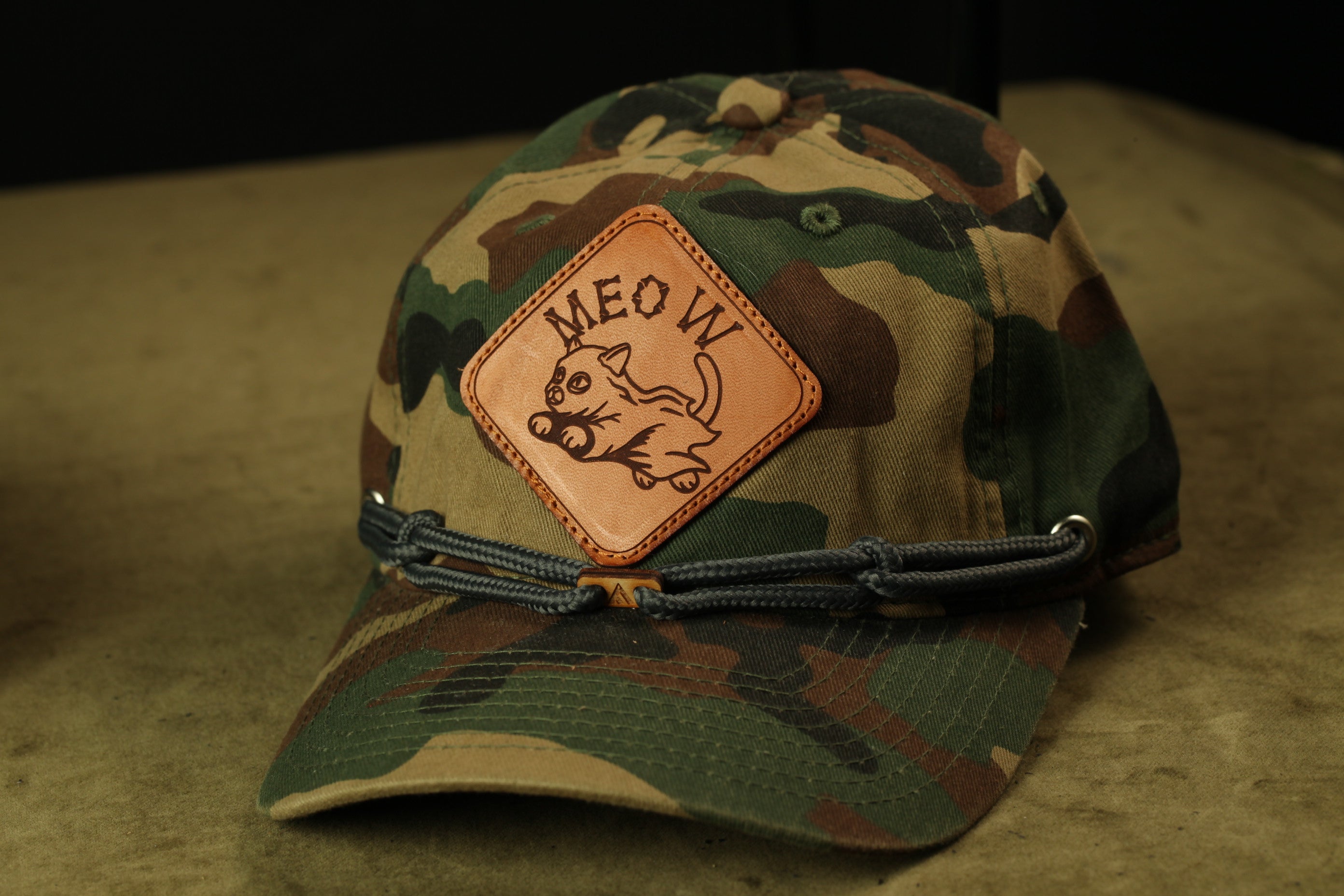 A camouflage baseball cap with a patch featuring a cartoon cat and the text "MEOW" printed on it.